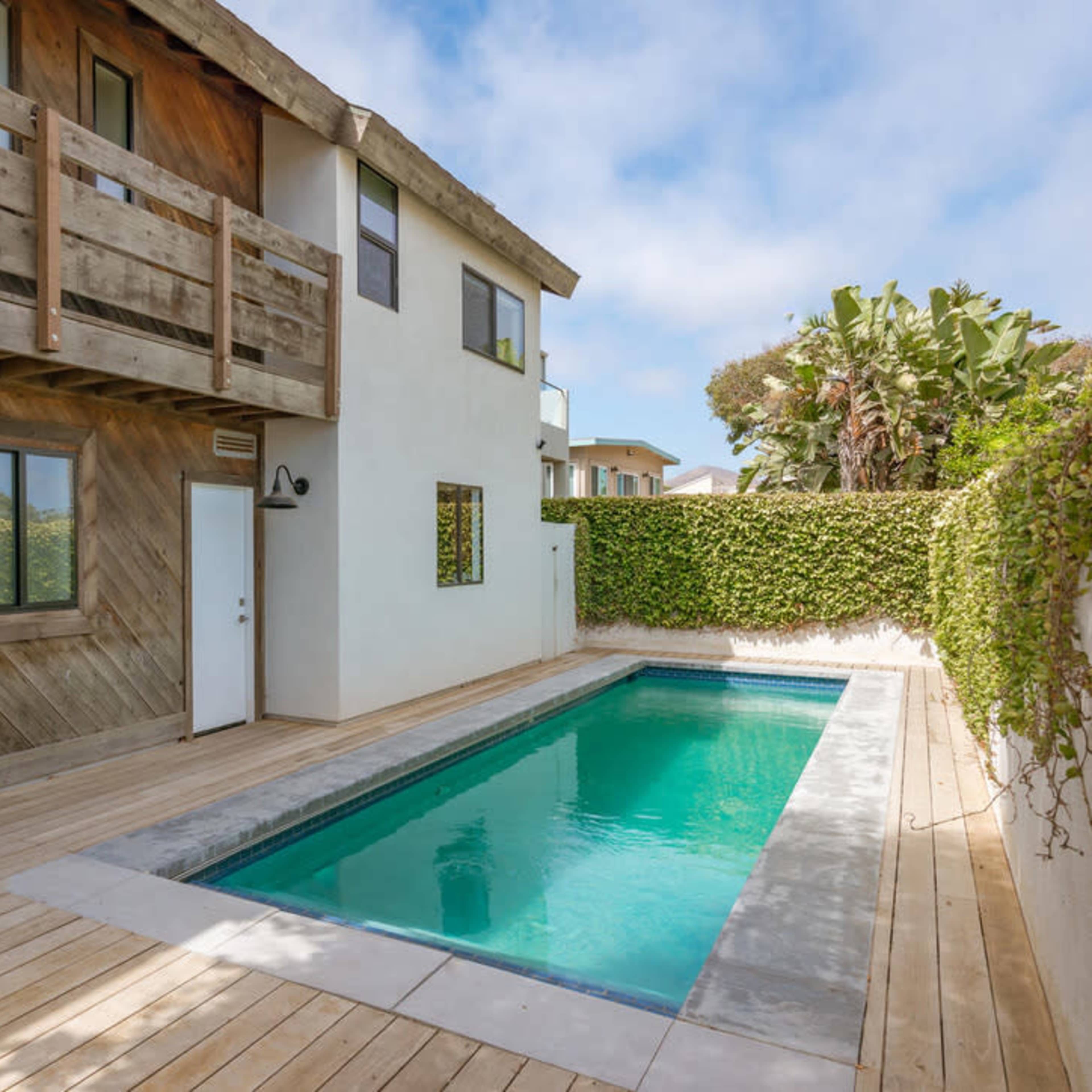 A modern house with a wooden deck and a rectangular swimming pool surrounded by greenery.