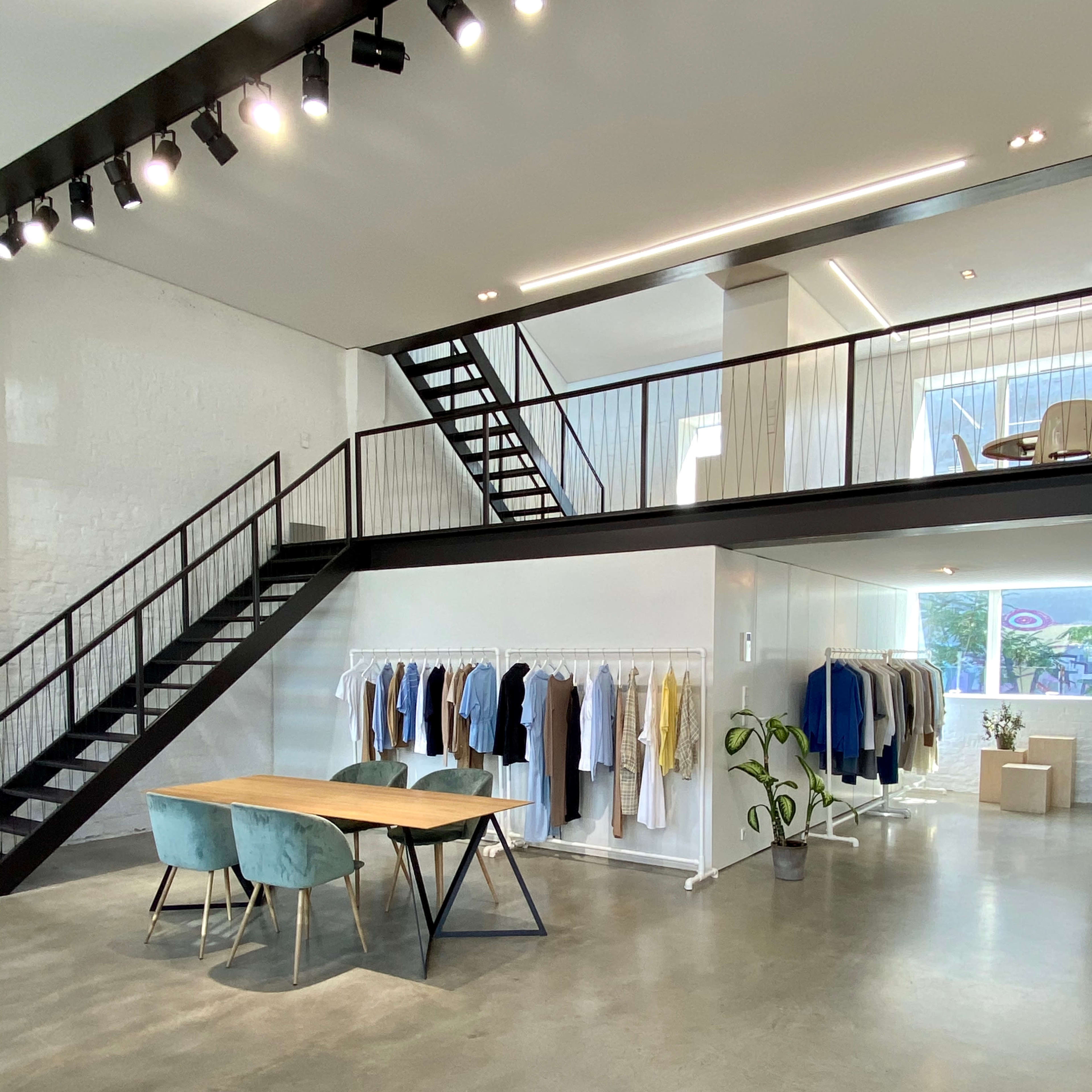 A modern clothing store features a wooden table with blue chairs, hanging garments on racks, and a staircase leading to an upper level.