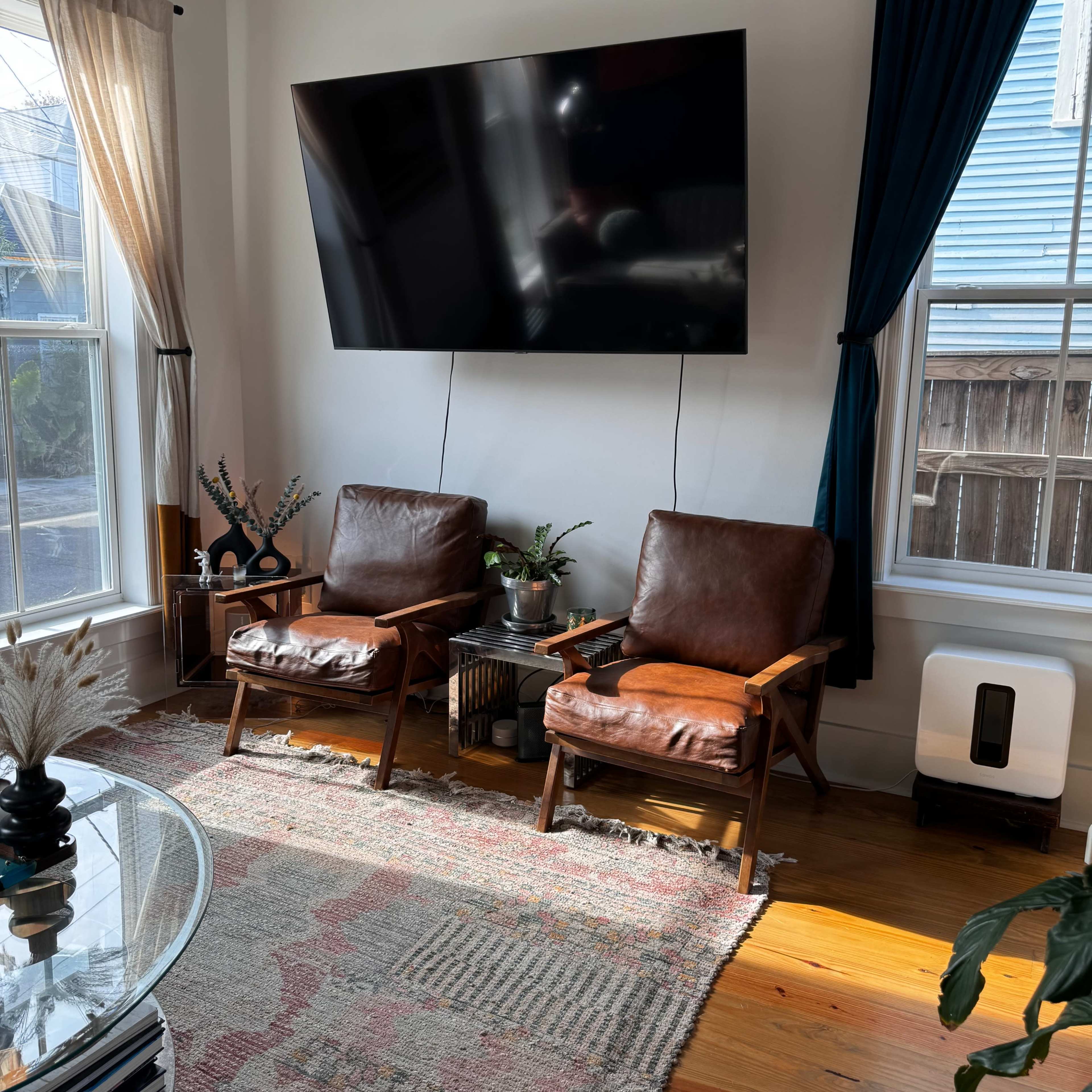 A living room with two brown leather chairs facing a large wall-mounted television, surrounded by windows with light curtains and a patterned rug on the floor.