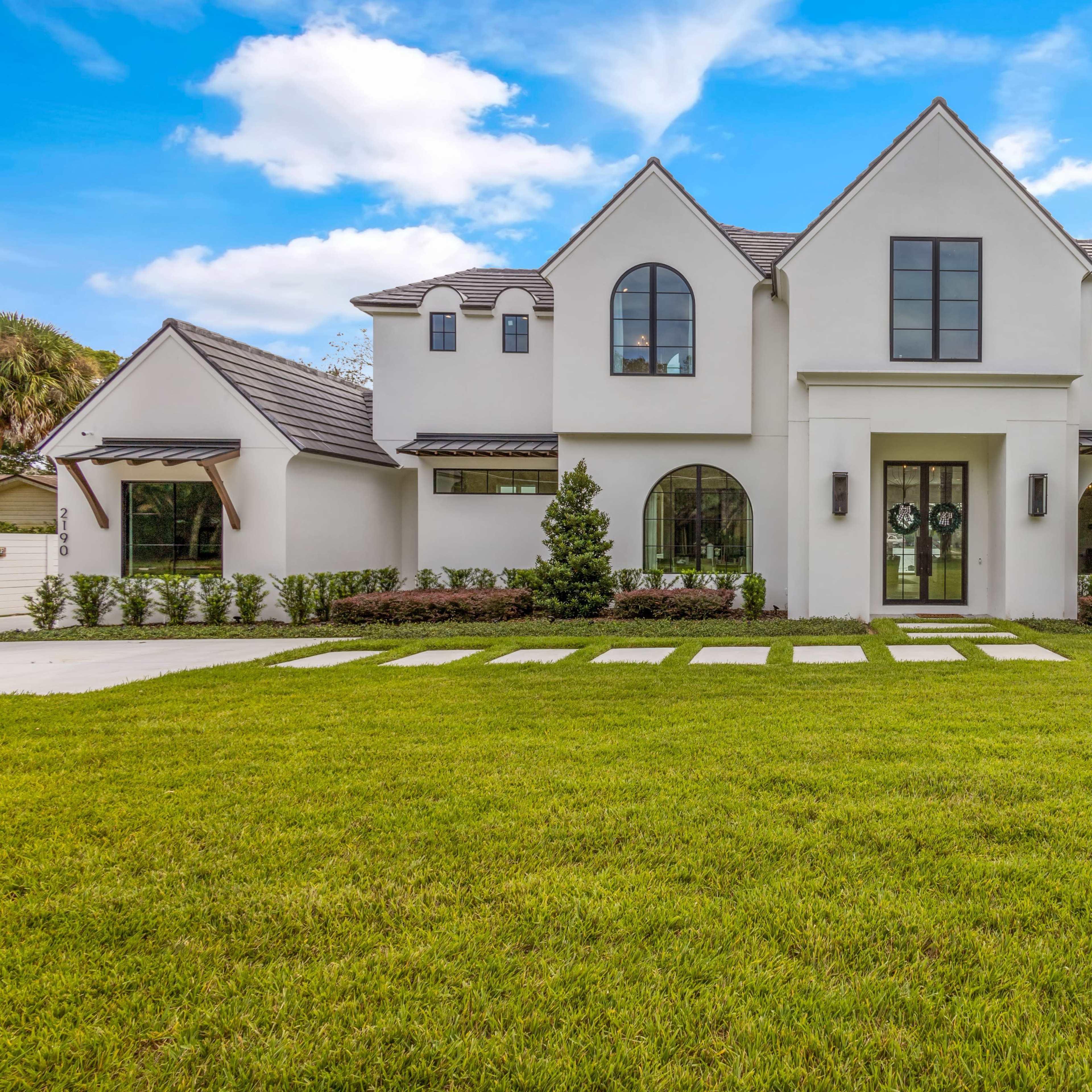 A two-story white house with a black roof features arched windows, a large front porch, and a manicured lawn with stone pathways.
