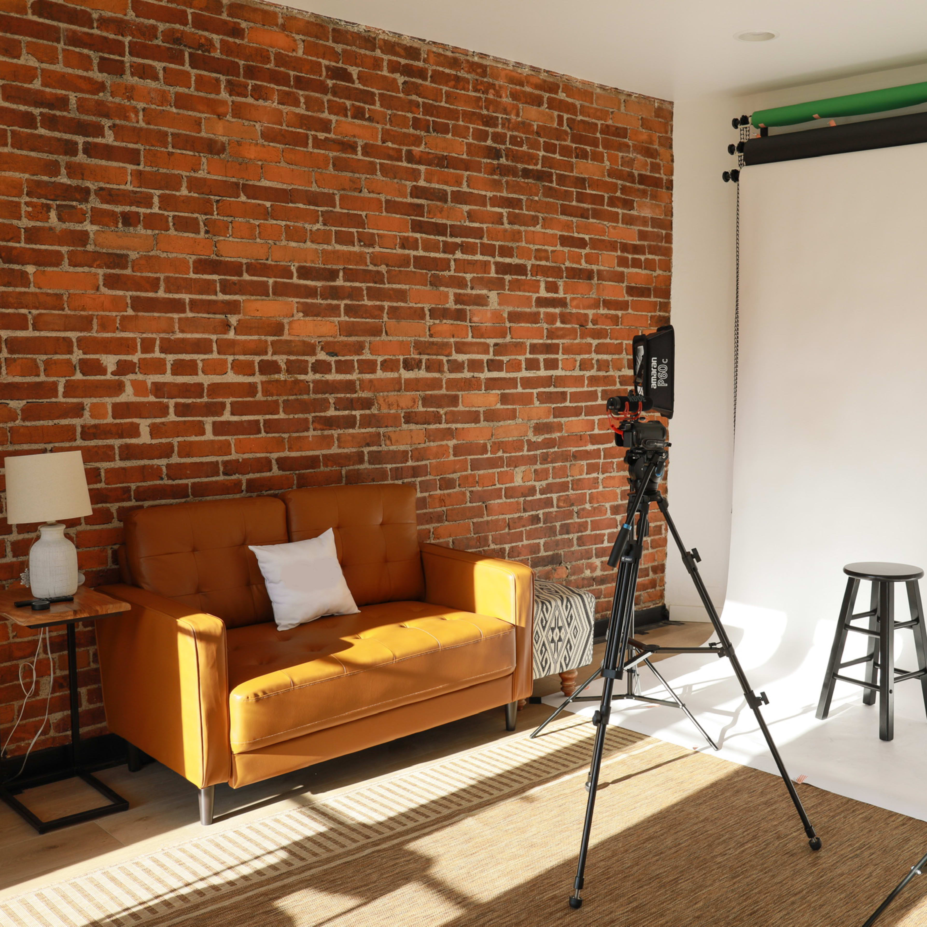 A photography studio with a brick wall backdrop, a brown sofa, a stool, and lighting equipment set up on a white backdrop.
