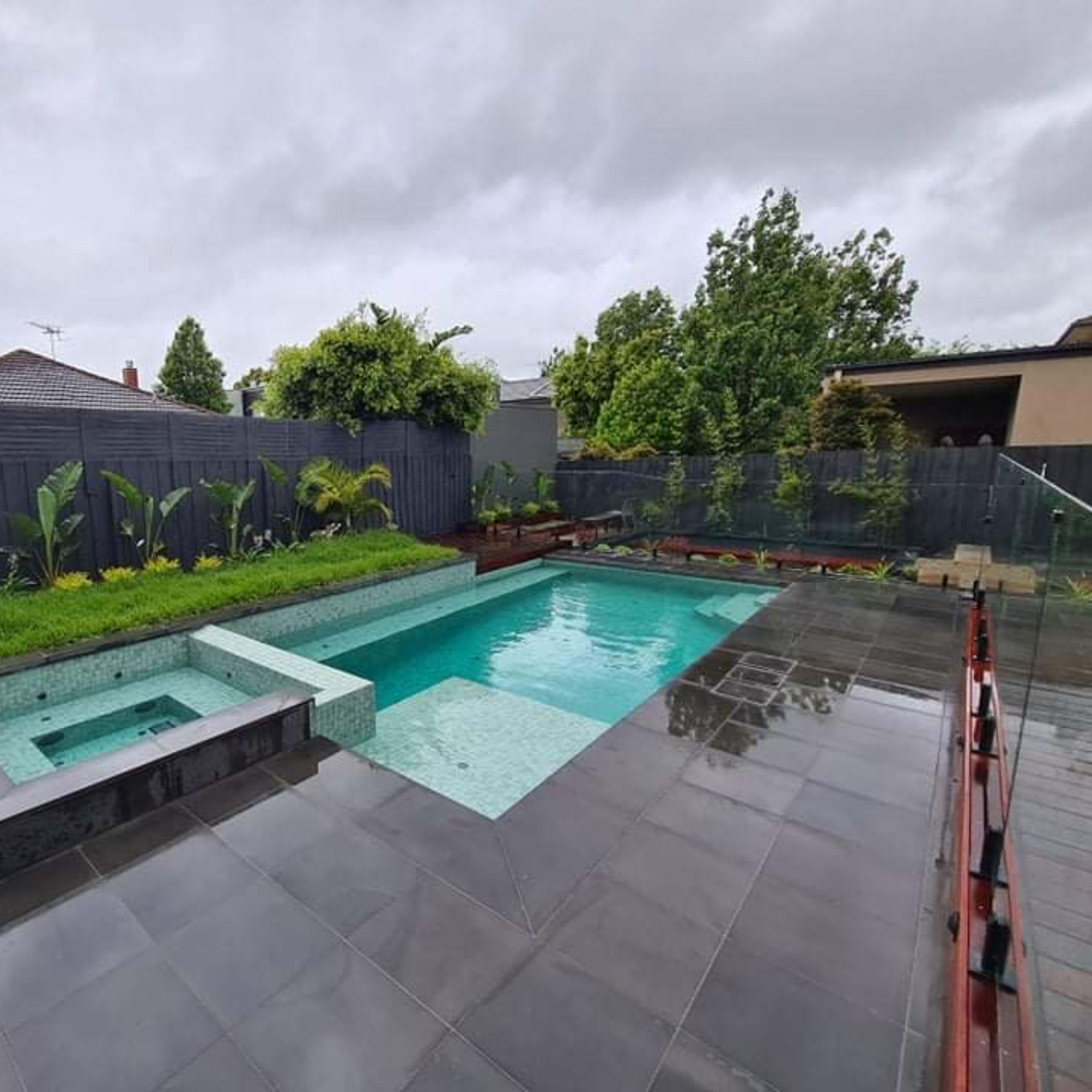 A modern backyard featuring a rectangular swimming pool, a spa area, and landscaped greenery surrounded by a wooden fence.