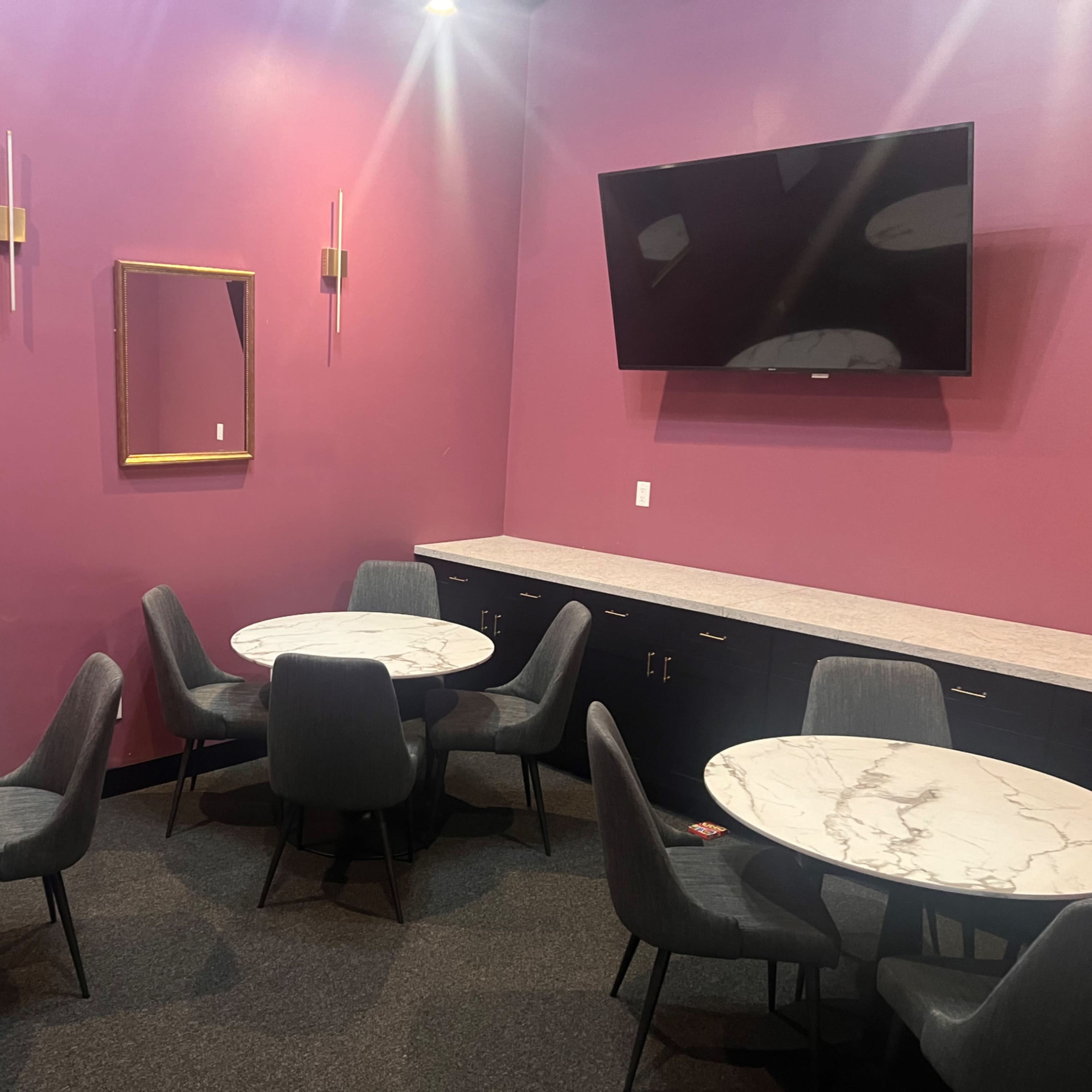 A small lounge area with several round marble tables and gray chairs against a purple wall, featuring a mirror and a television on the wall.