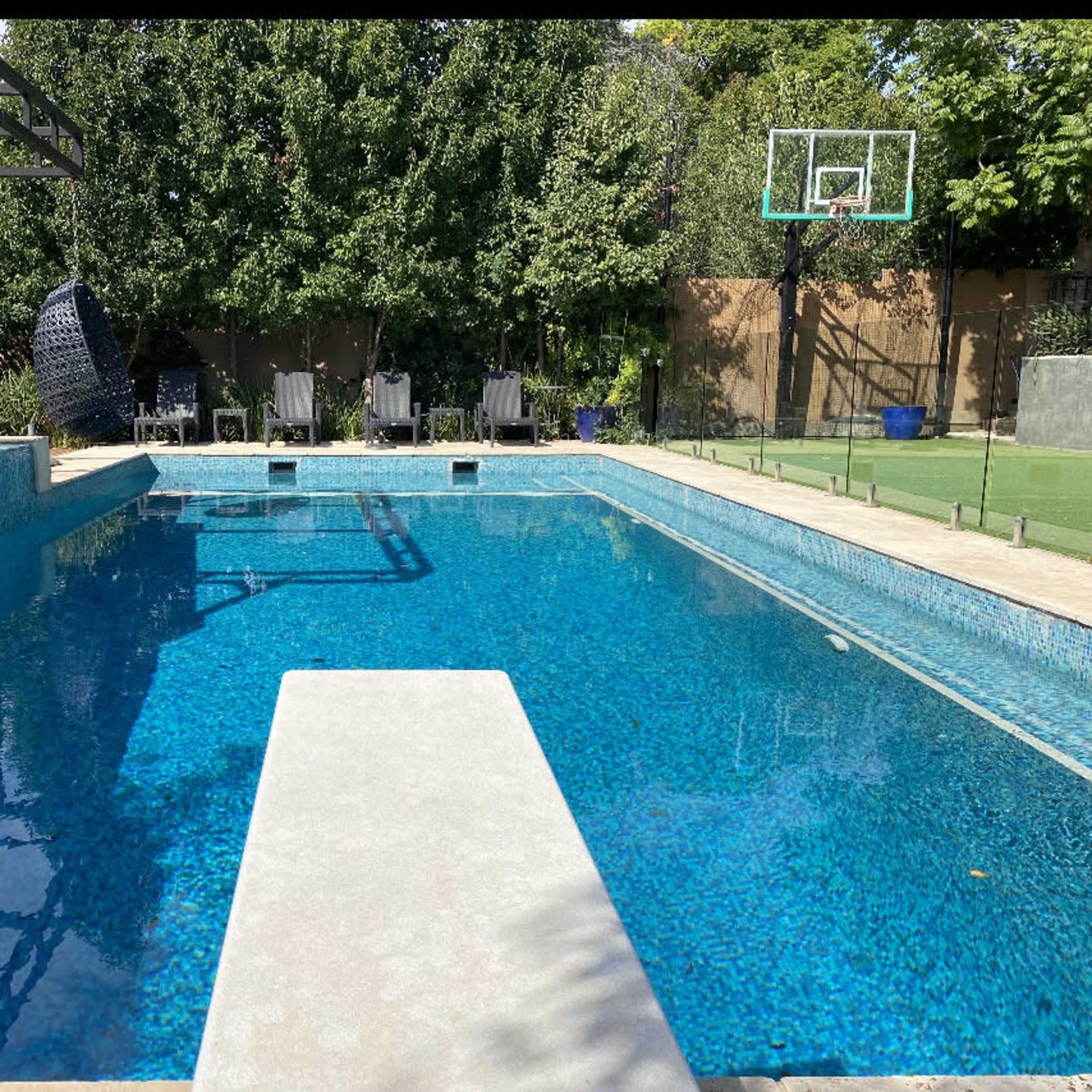 A rectangular swimming pool with a diving board, surrounded by lounge chairs and a basketball hoop in a landscaped backyard.