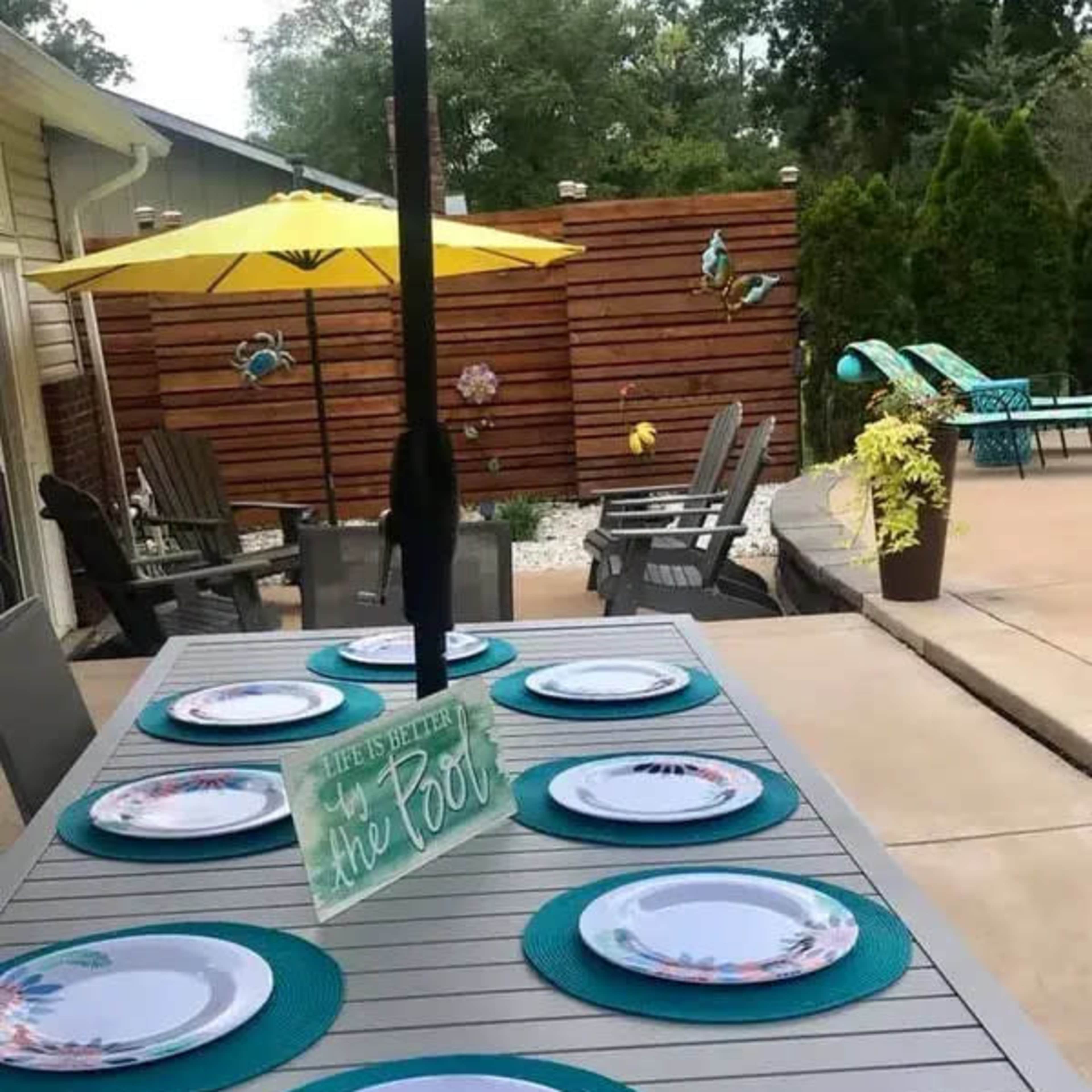 A dining table is set with plates and teal placemats under an umbrella, adjacent to a backyard area featuring an umbrella and decorative elements.