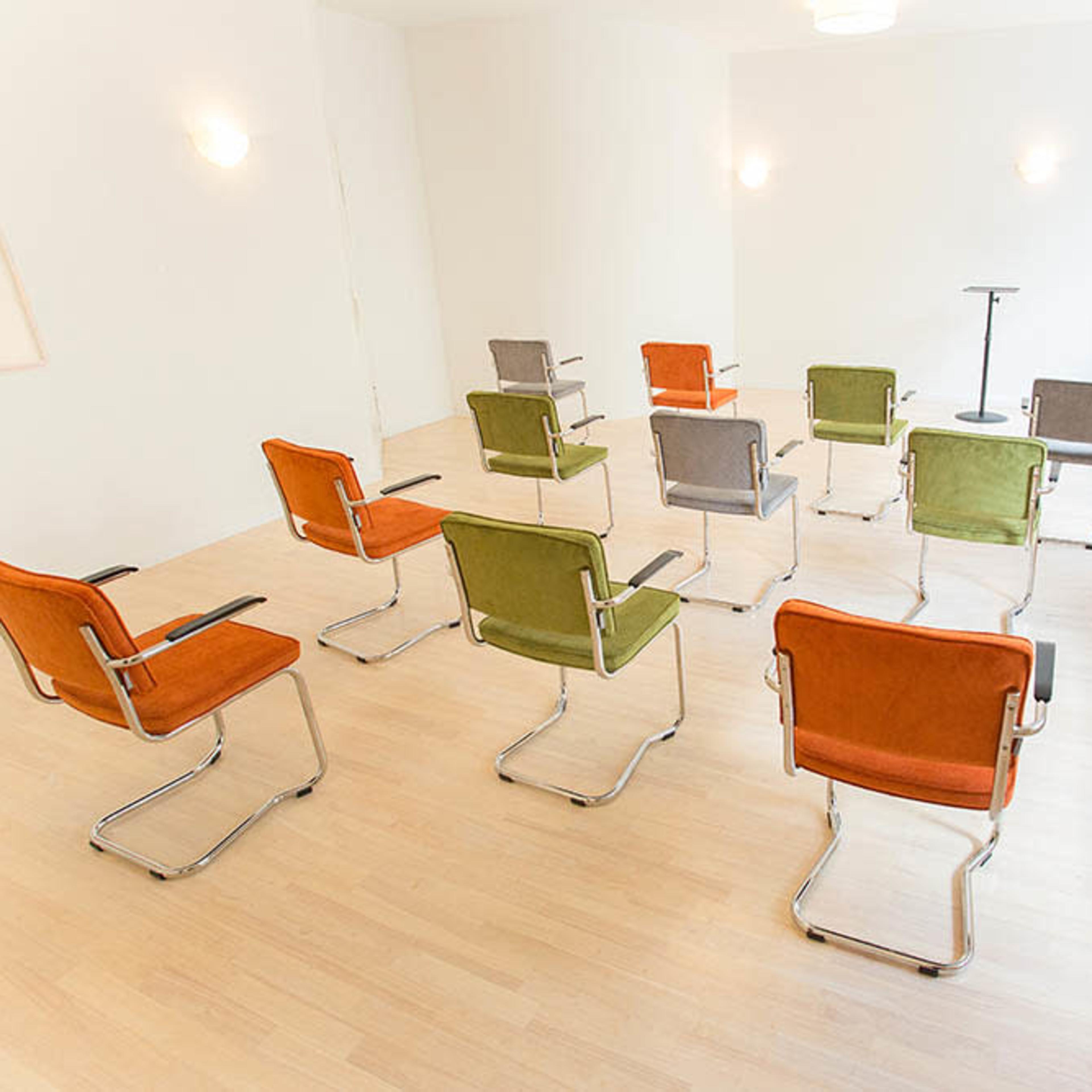 A sparse room features a circular arrangement of ten colorful chairs with a small podium at the center.