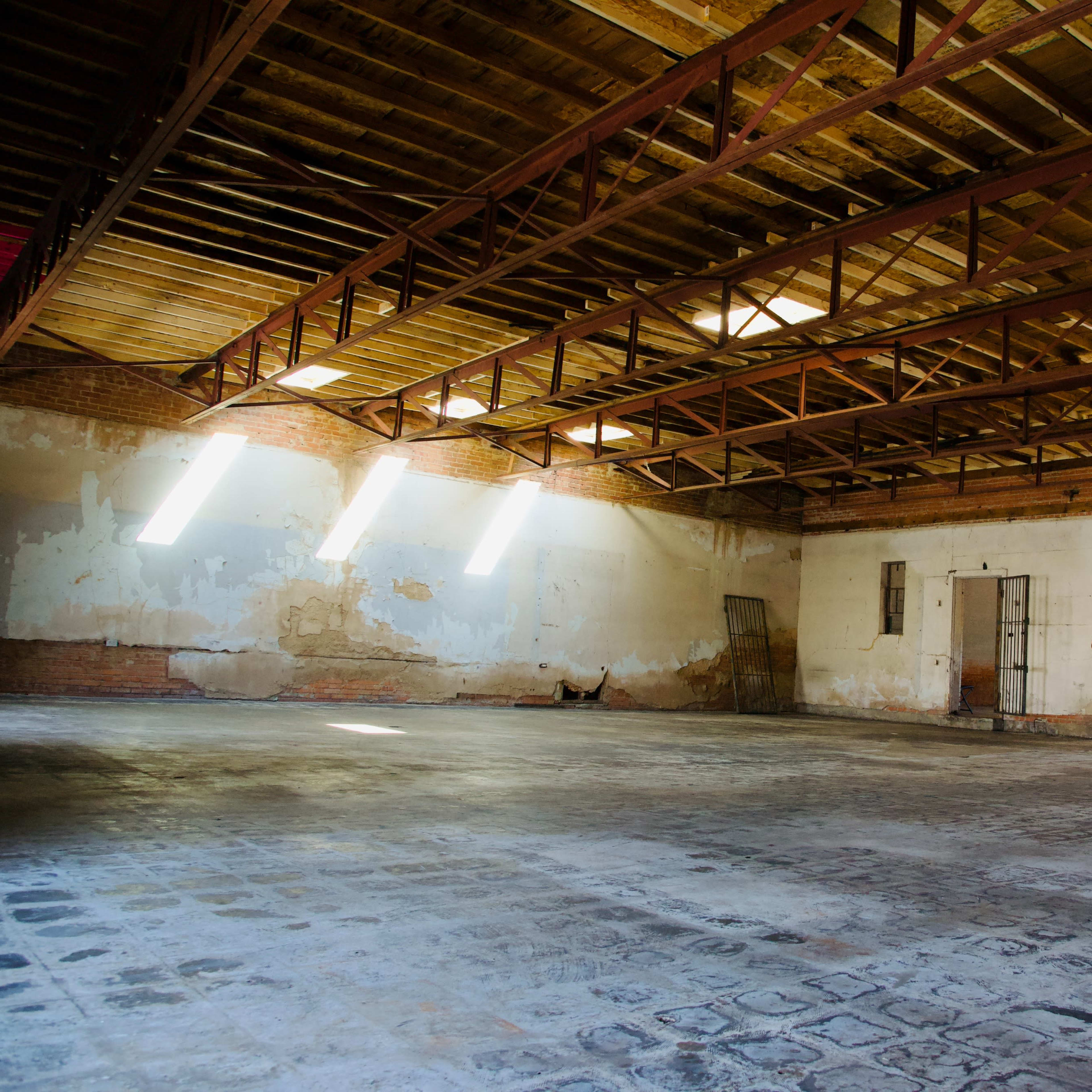 A spacious, empty industrial room with exposed wooden beams and sunlight filtering through large windows onto a bare floor.
