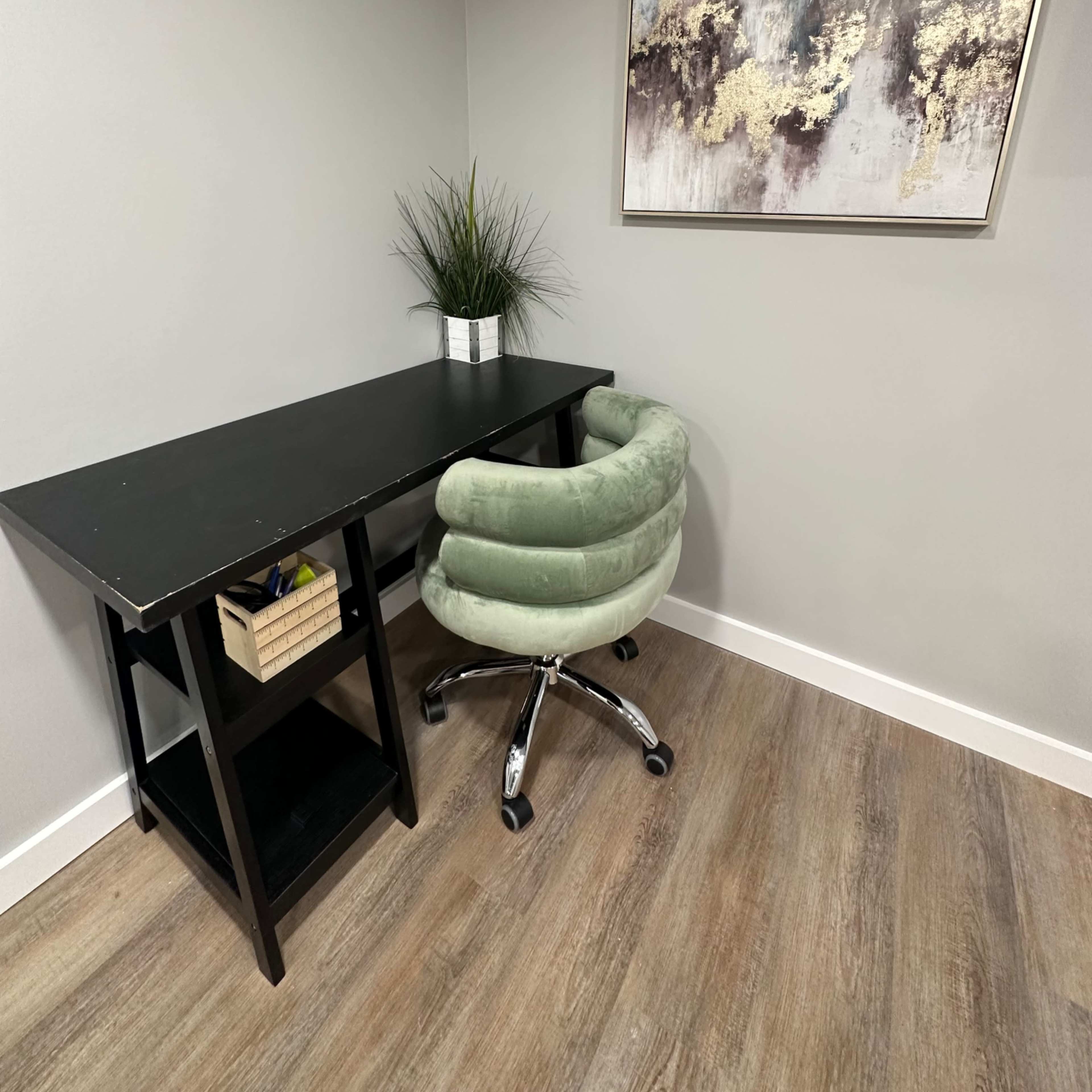 A minimalist workspace features a black desk with a green swivel chair and a potted plant, against a wall adorned with abstract art.