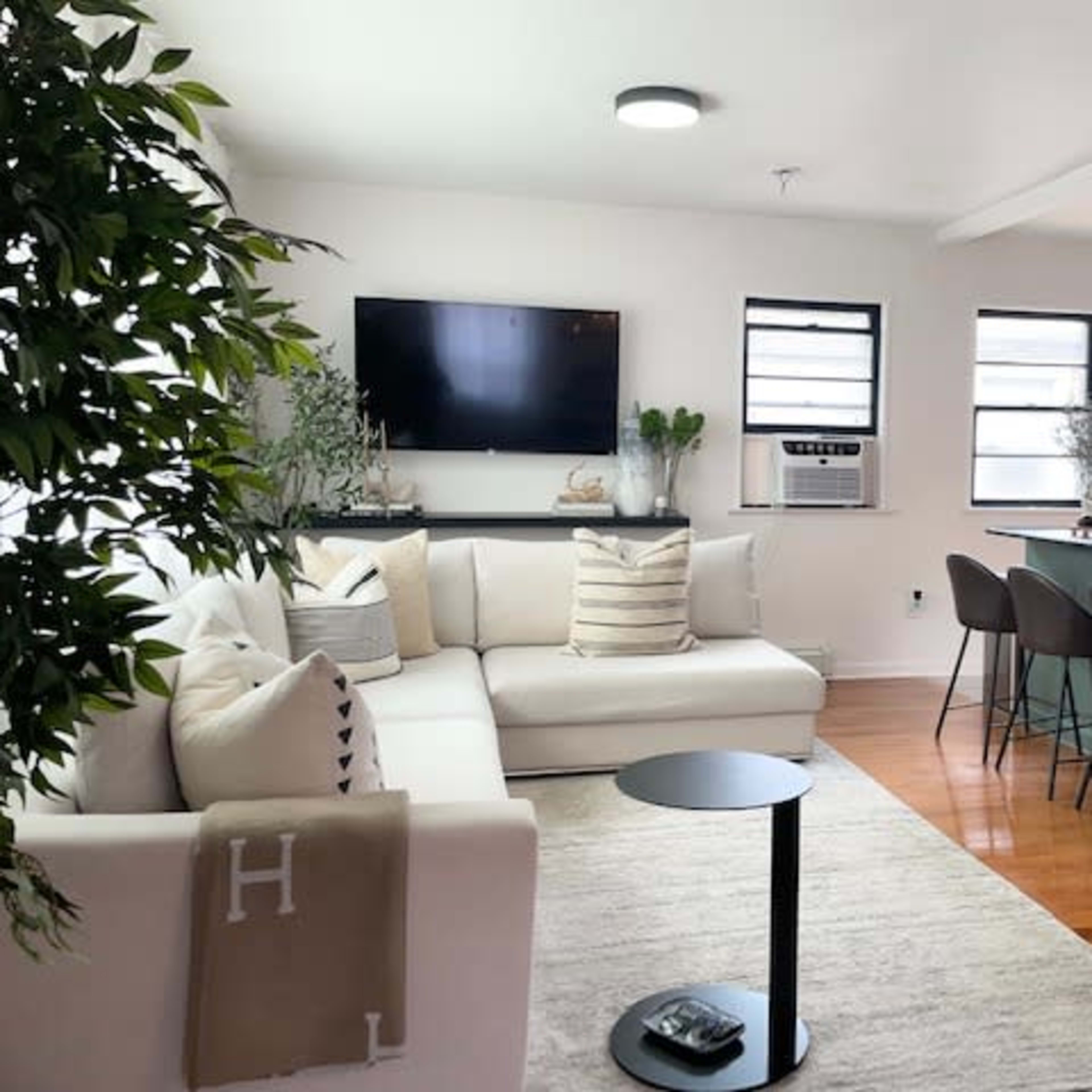A bright living room with a white sectional sofa, a small black side table, a television mounted on the wall, and a kitchen counter with bar stools in the background.