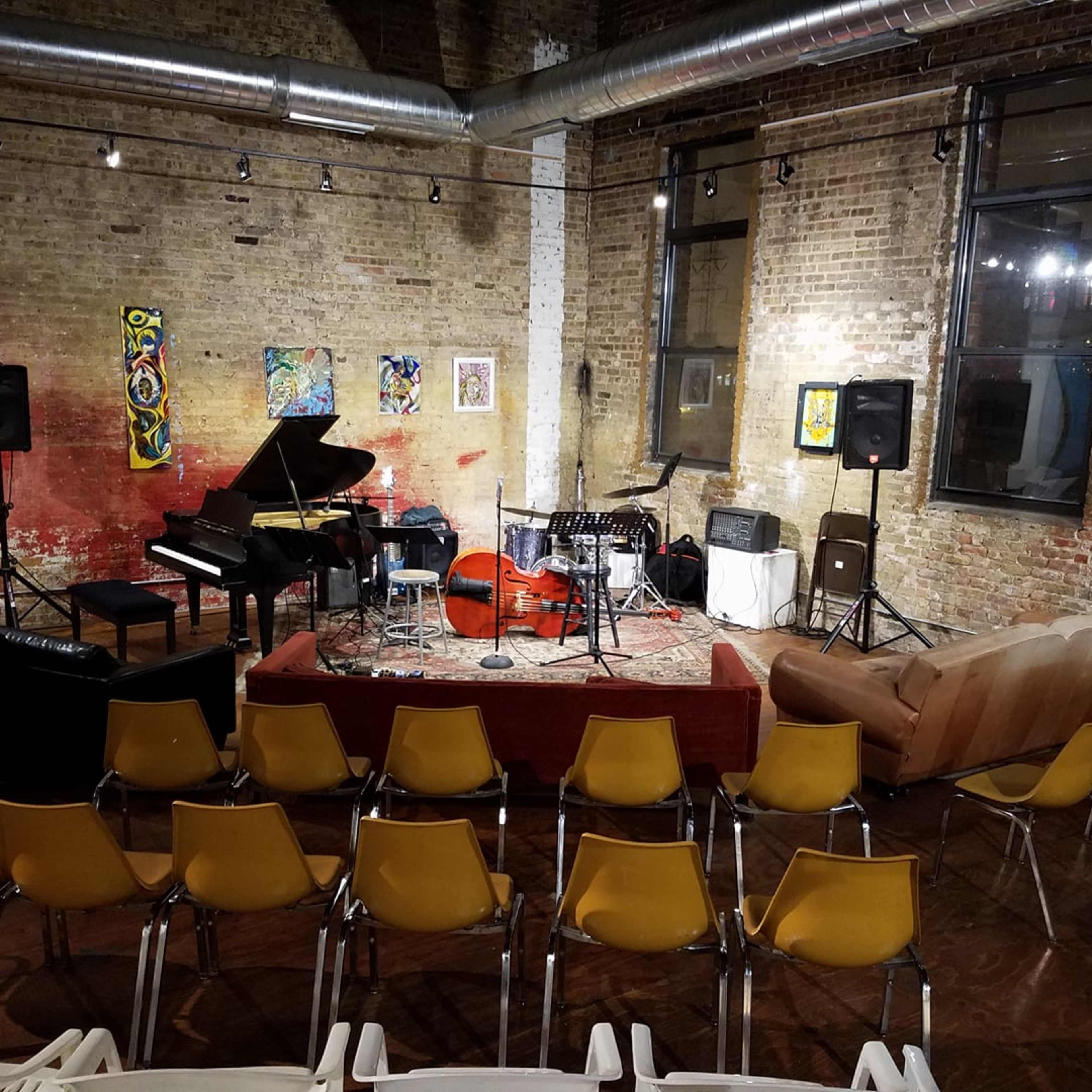 A small performance space features a piano, drum set, and string bass, arranged on a rug in front of chairs and a couch.