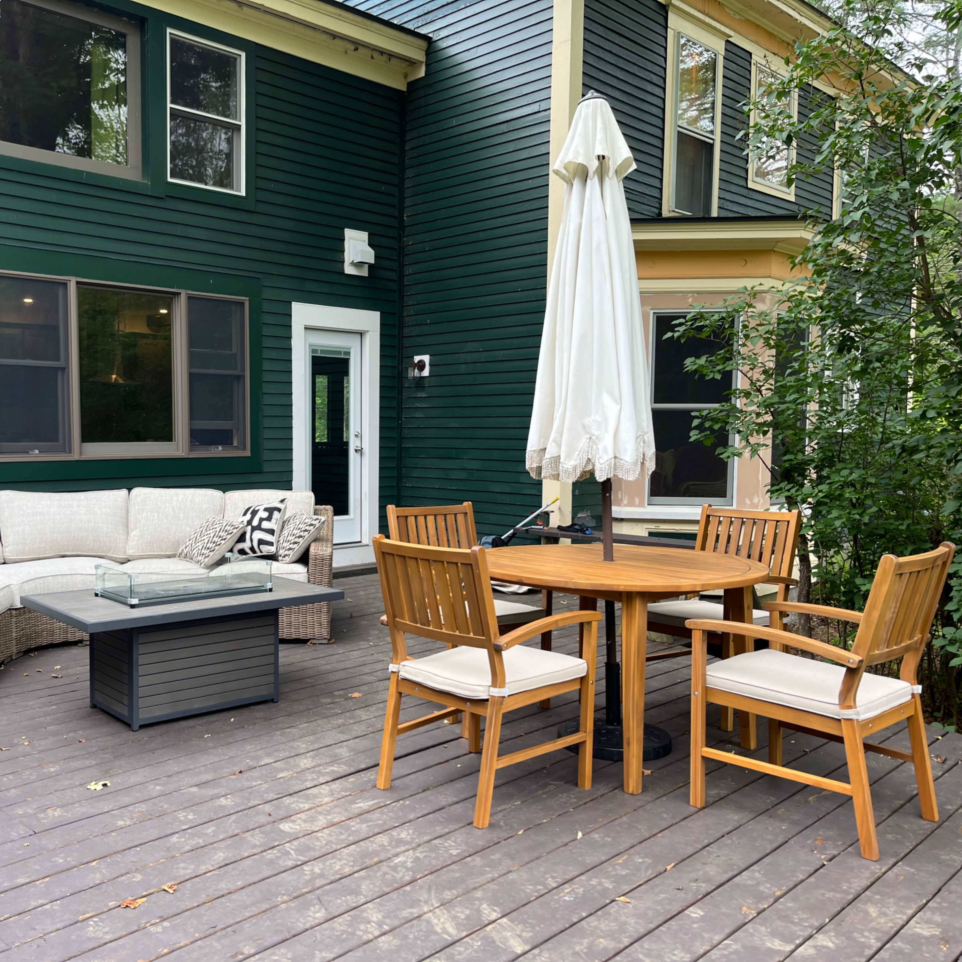 A cozy outdoor patio features a sectional sofa, a circular dining table with four chairs, and a large umbrella, all set against a green wooden house.