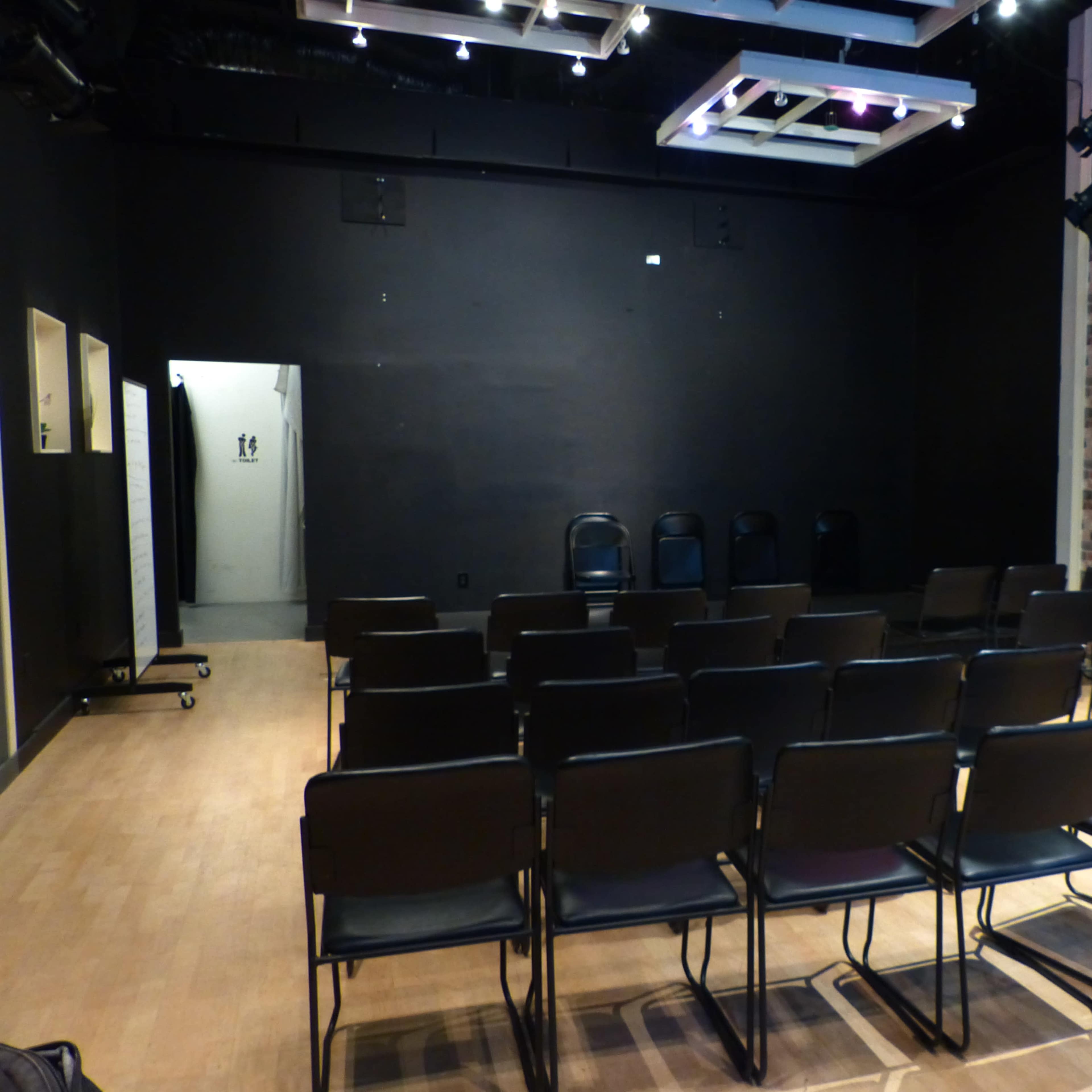 A small theater or meeting room with rows of black chairs facing a blank wall, and a doorway leading to a restroom area.