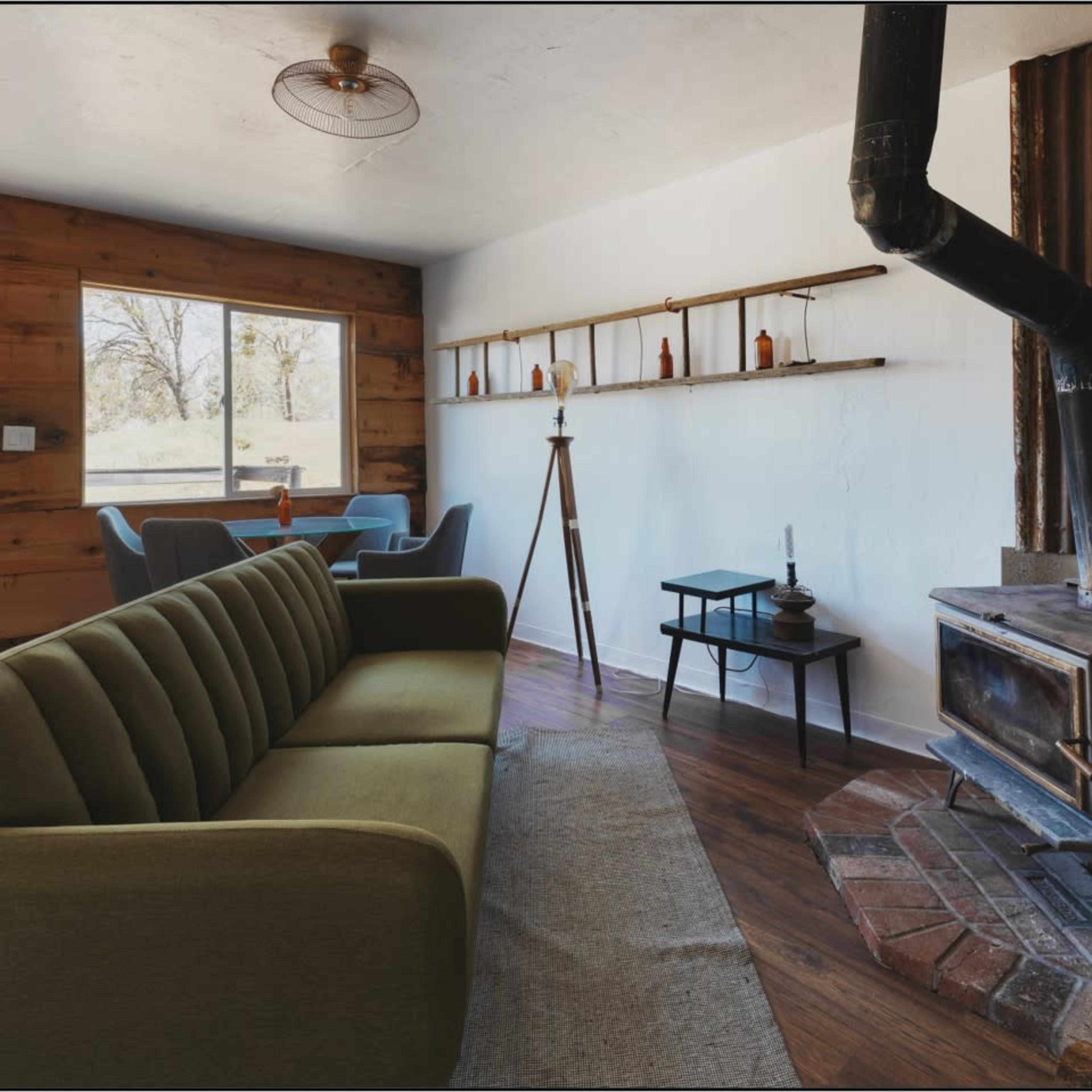 The room features a green sofa, a wooden dining area, and a metal stove against a rustic backdrop.