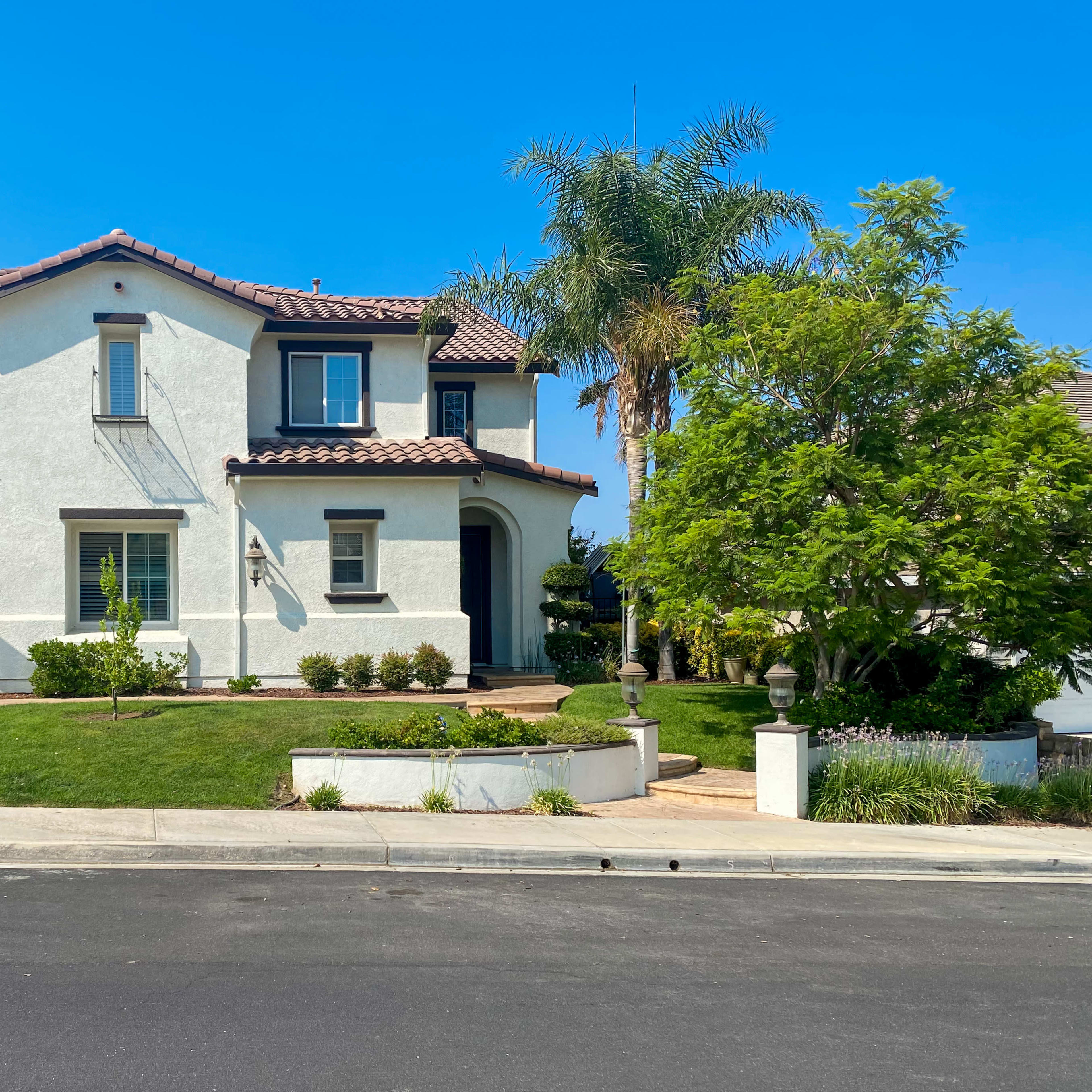 A two-story white house with a tile roof and a front yard featuring green grass and shrubs is located on a residential street.