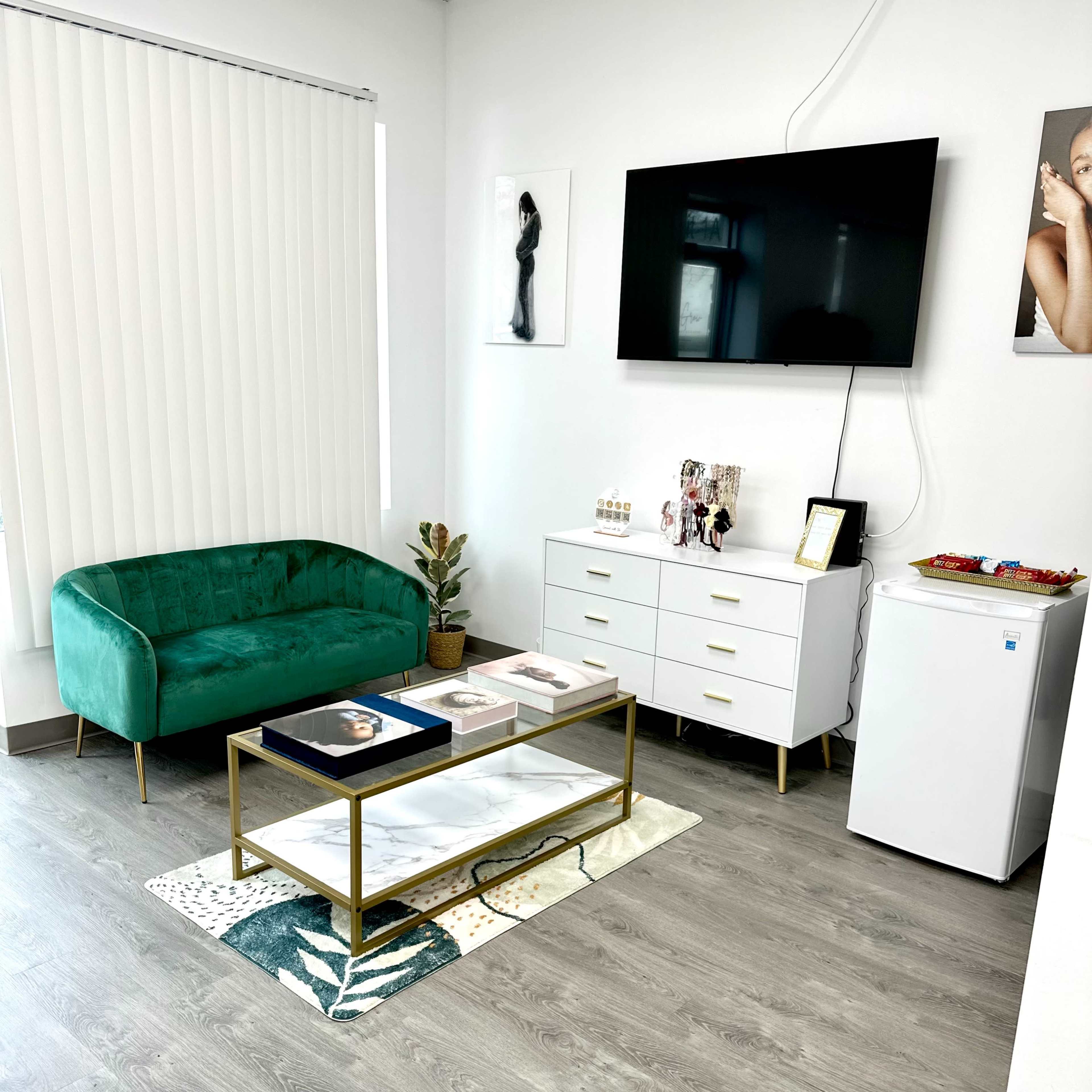 A modern waiting area with a green velvet couch, a wall-mounted television, a white dresser, and a small refrigerator, all arranged on a light wood floor.