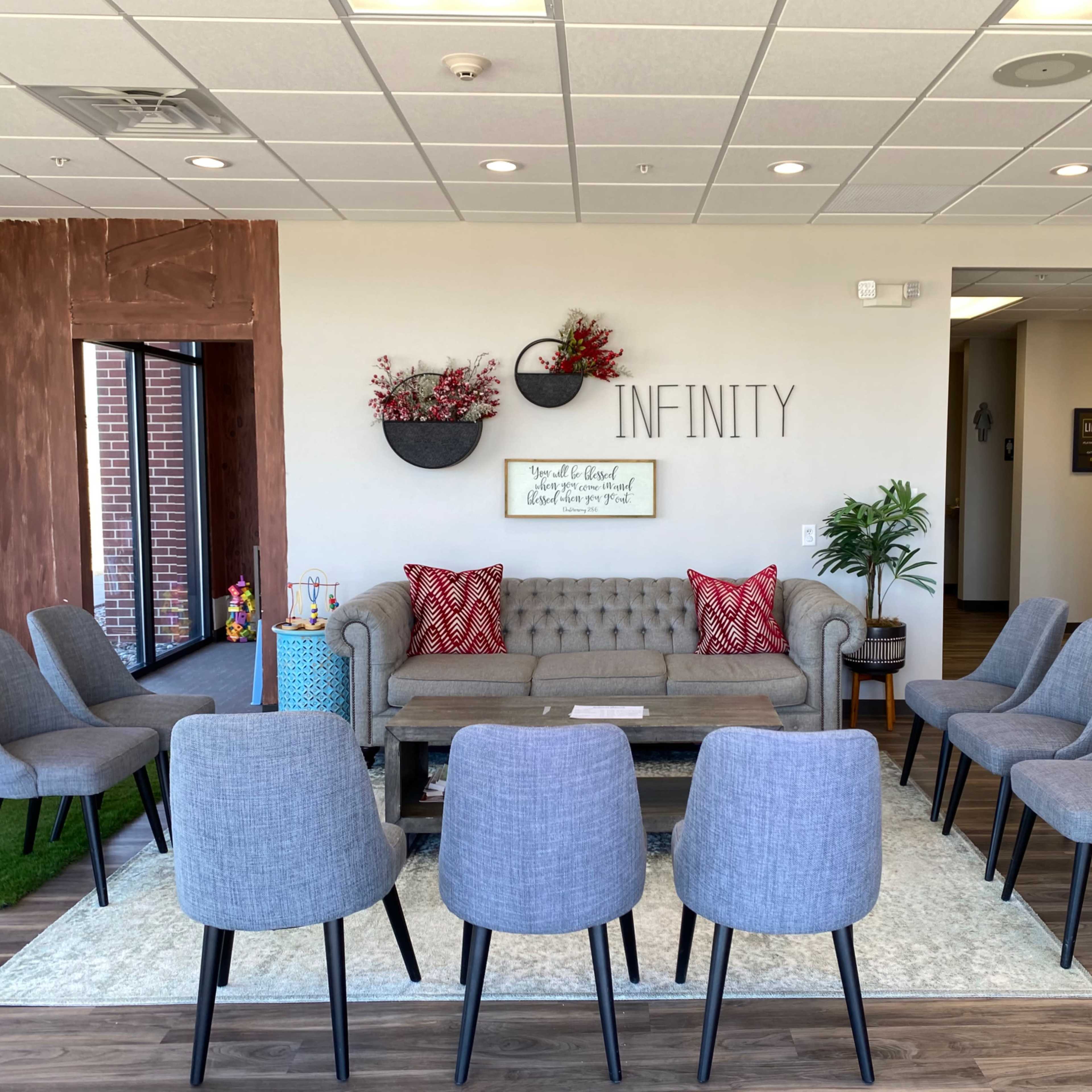 A waiting area features a gray tufted sofa and six chairs arranged in a circle on a light rug, with wall decorations and a plant in the corner.