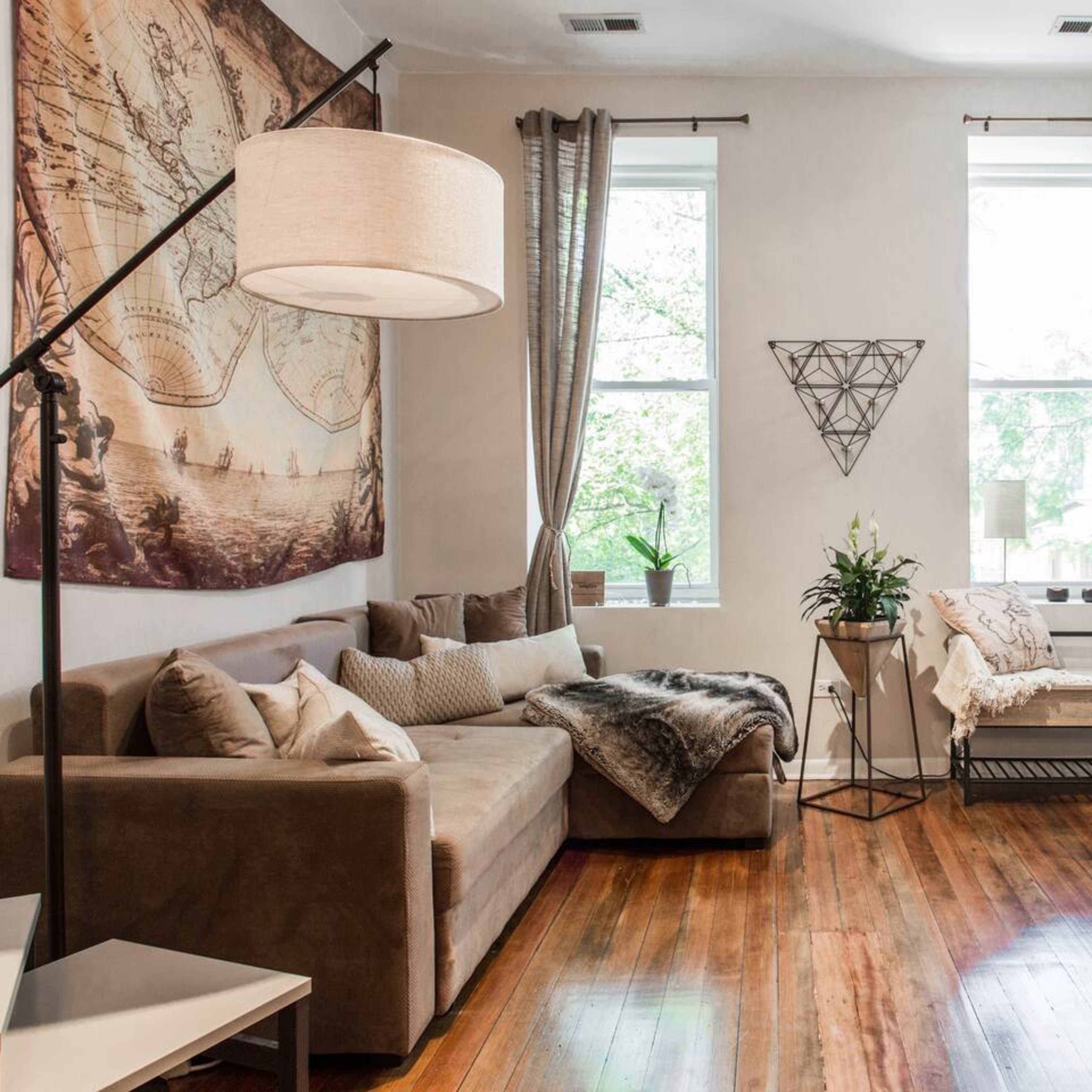 The room features a sofa with cushions, a light fixture, a plant in a corner, and large windows with curtains.