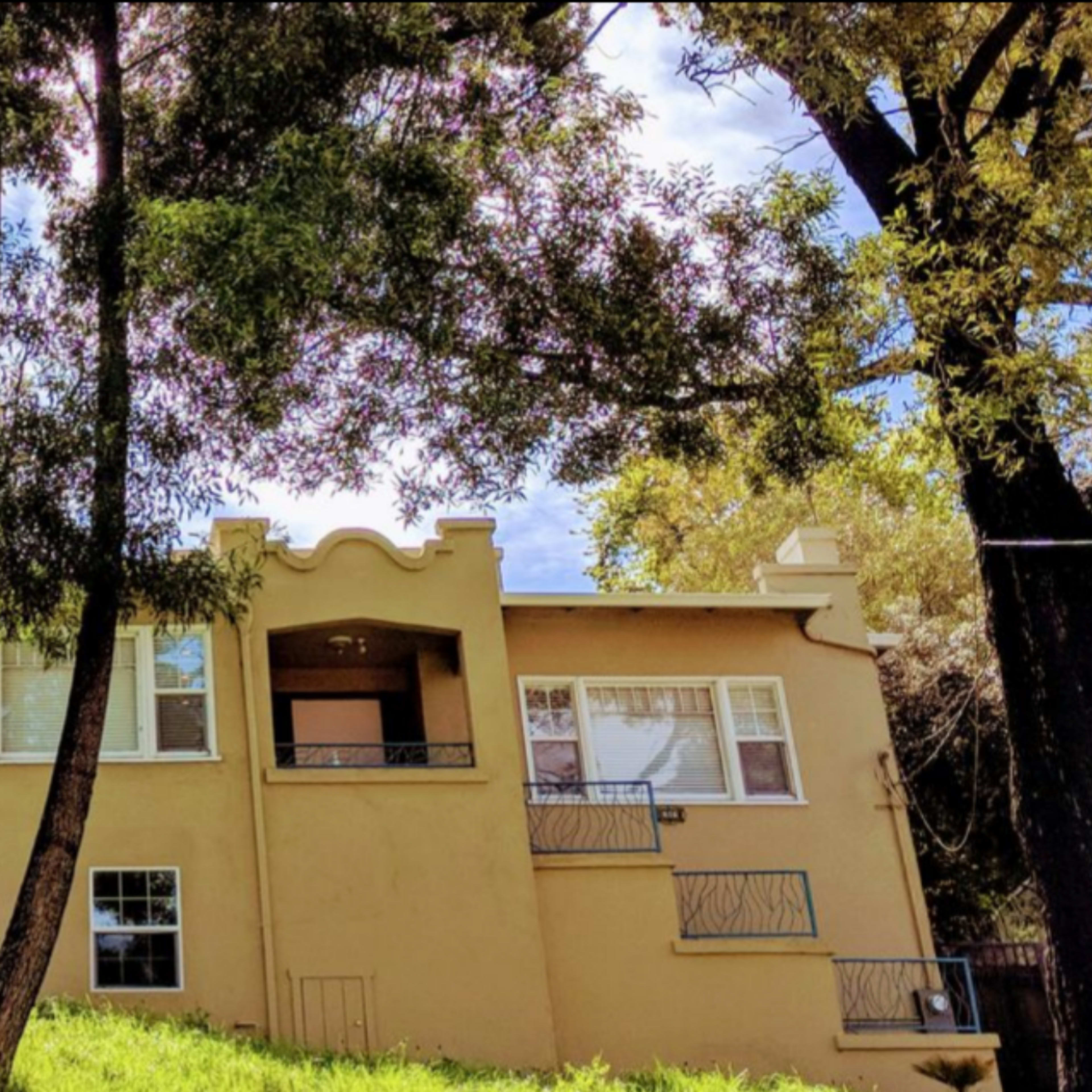A tan house with large windows and decorative rooflines sits on a grassy slope, surrounded by tall trees.