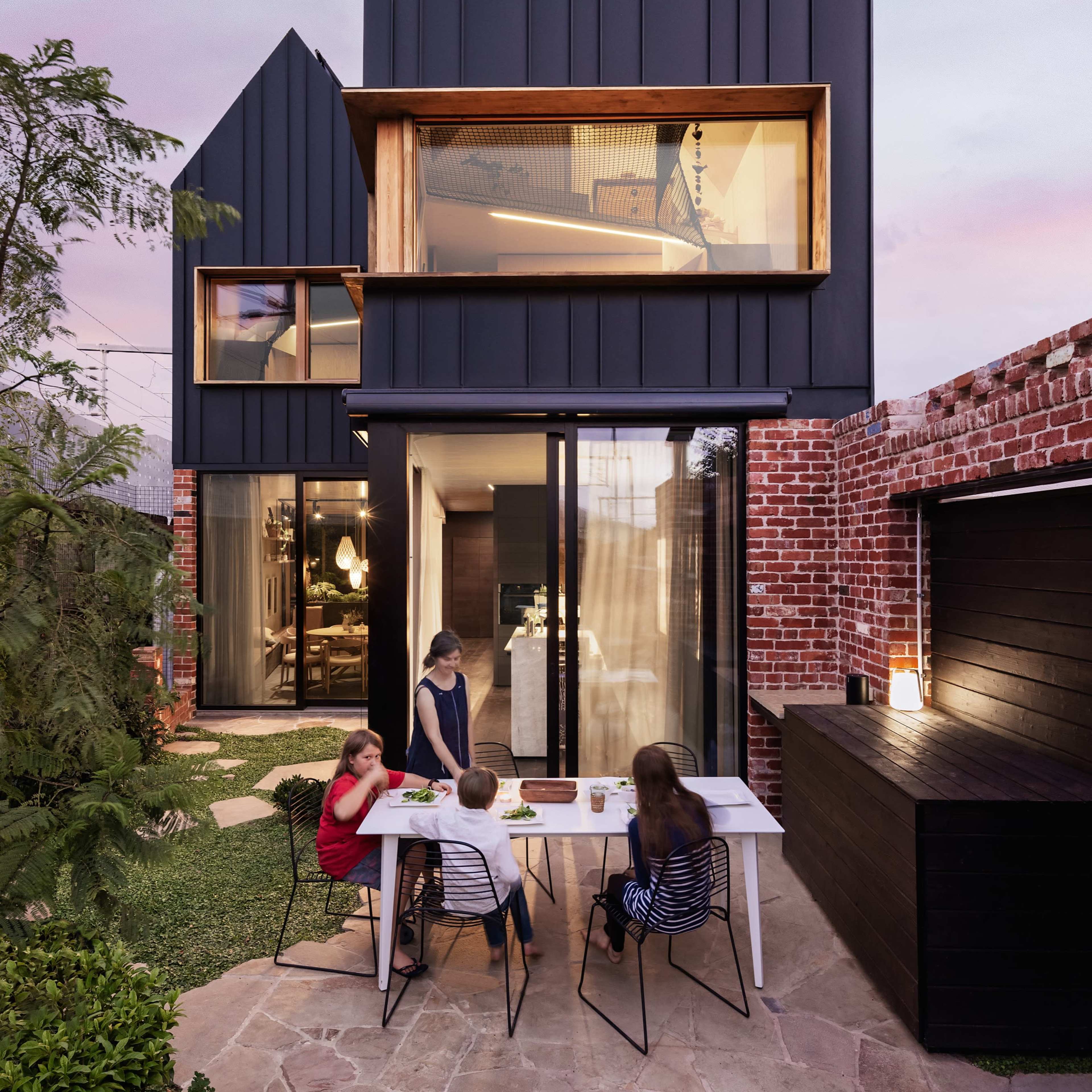 A modern black-clad house features large windows and angular roofs, as a family gathers around a table outdoors at dusk.