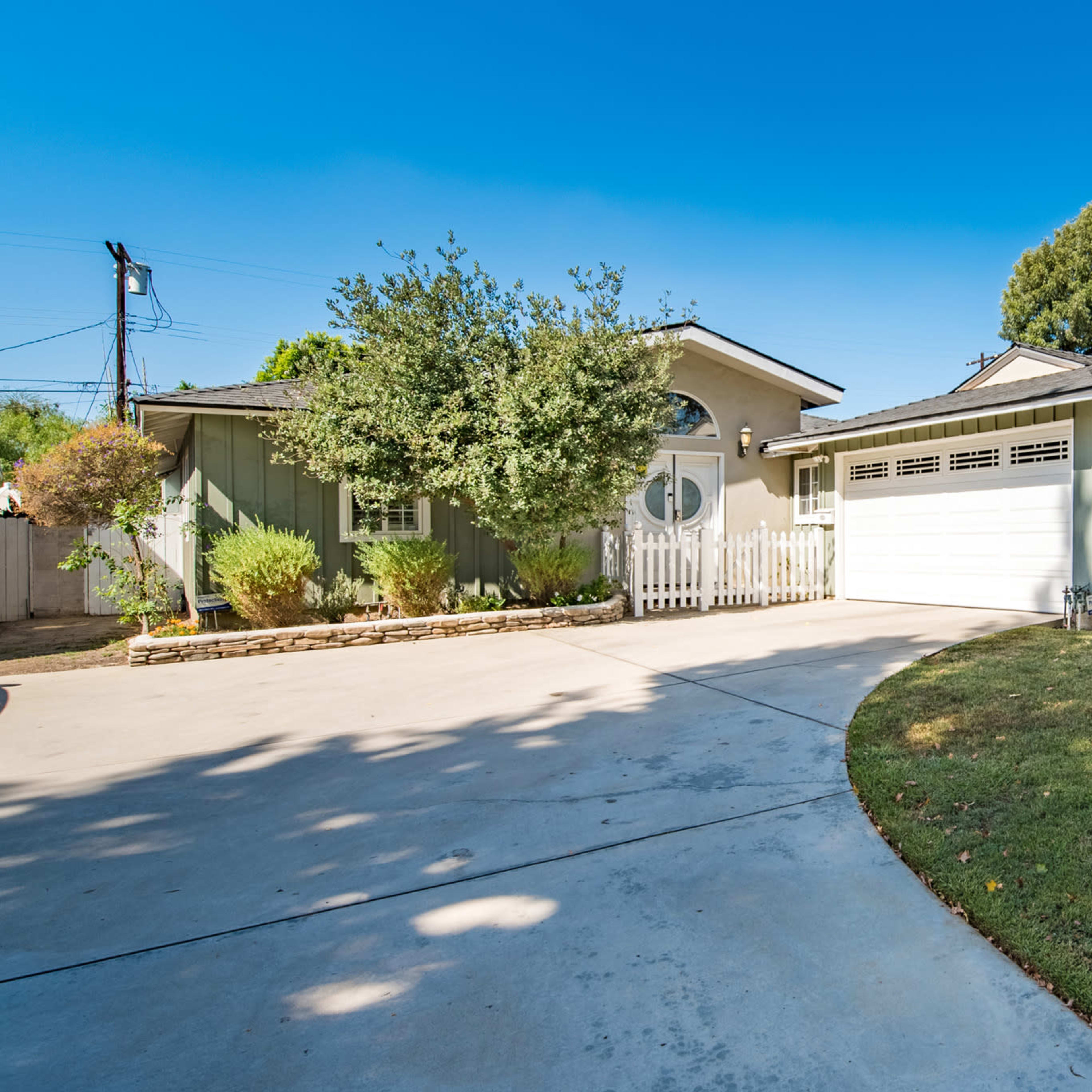 A single-story green house with a white picket fence and a spacious driveway is set against a clear blue sky.