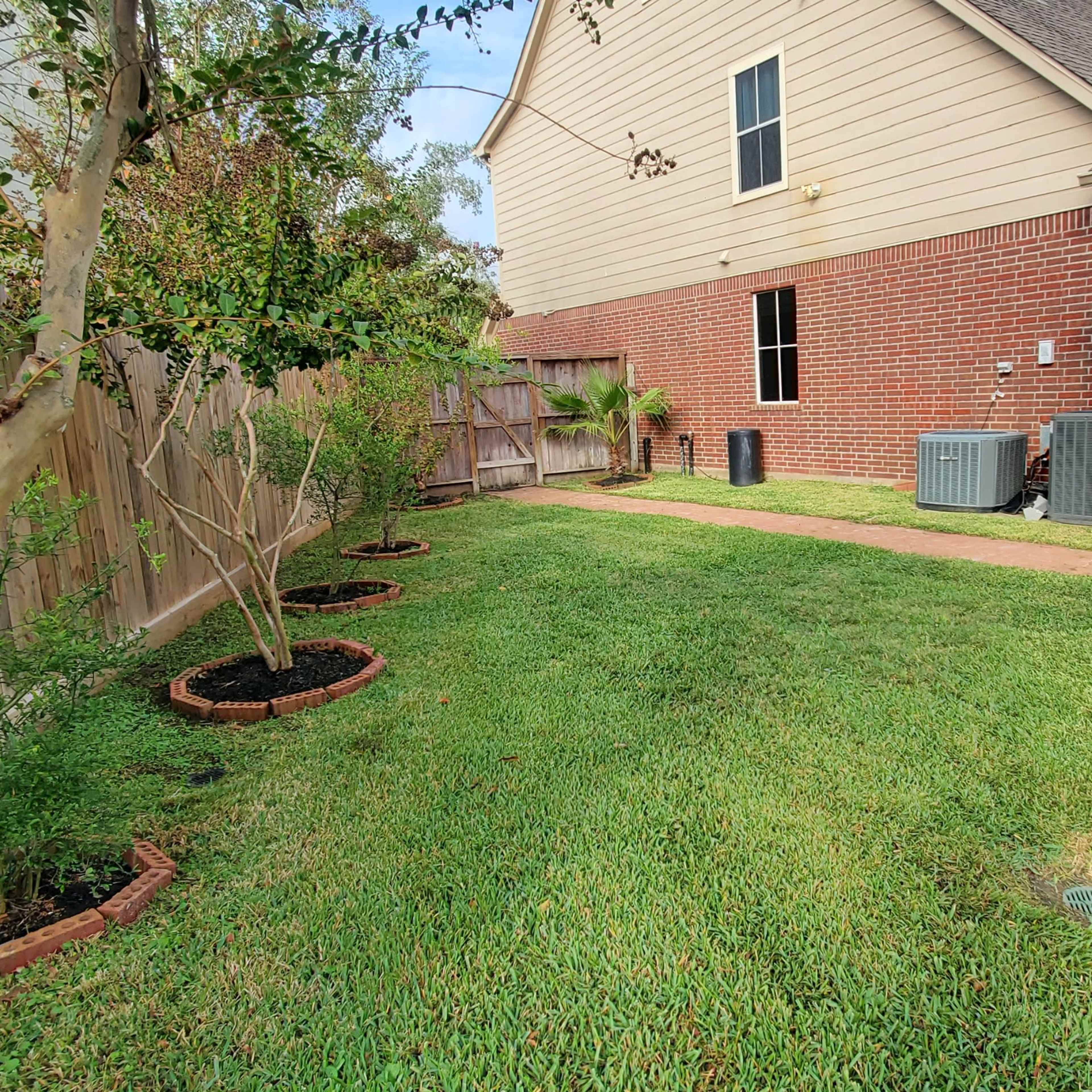 A neatly landscaped backyard with grassy areas, decorative plant beds, a small storage shed, and air conditioning units along a brick wall.