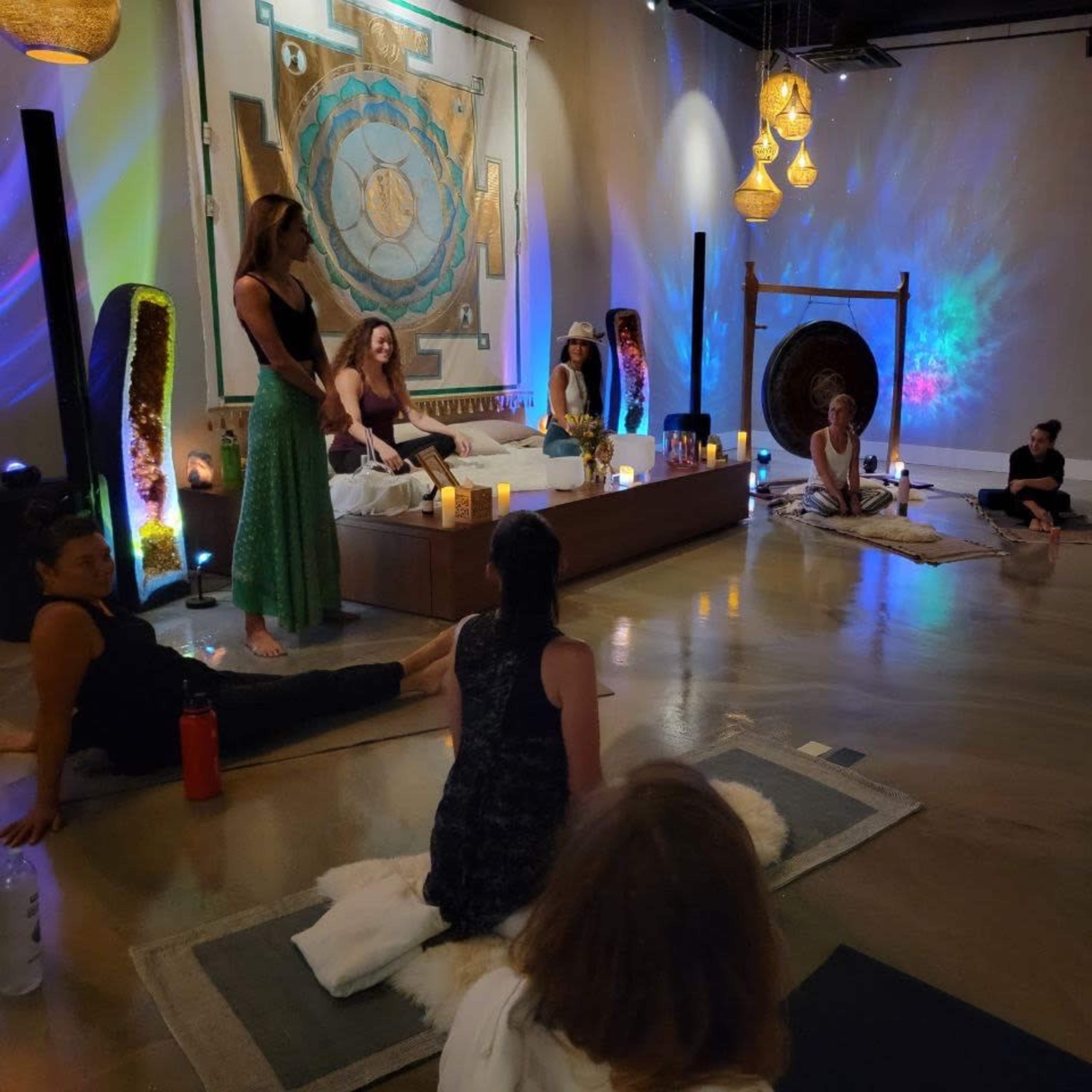 A group of people sit on the floor in a dimly lit room arranged for a wellness or meditation session, with colorful lights and decorative elements in the background.