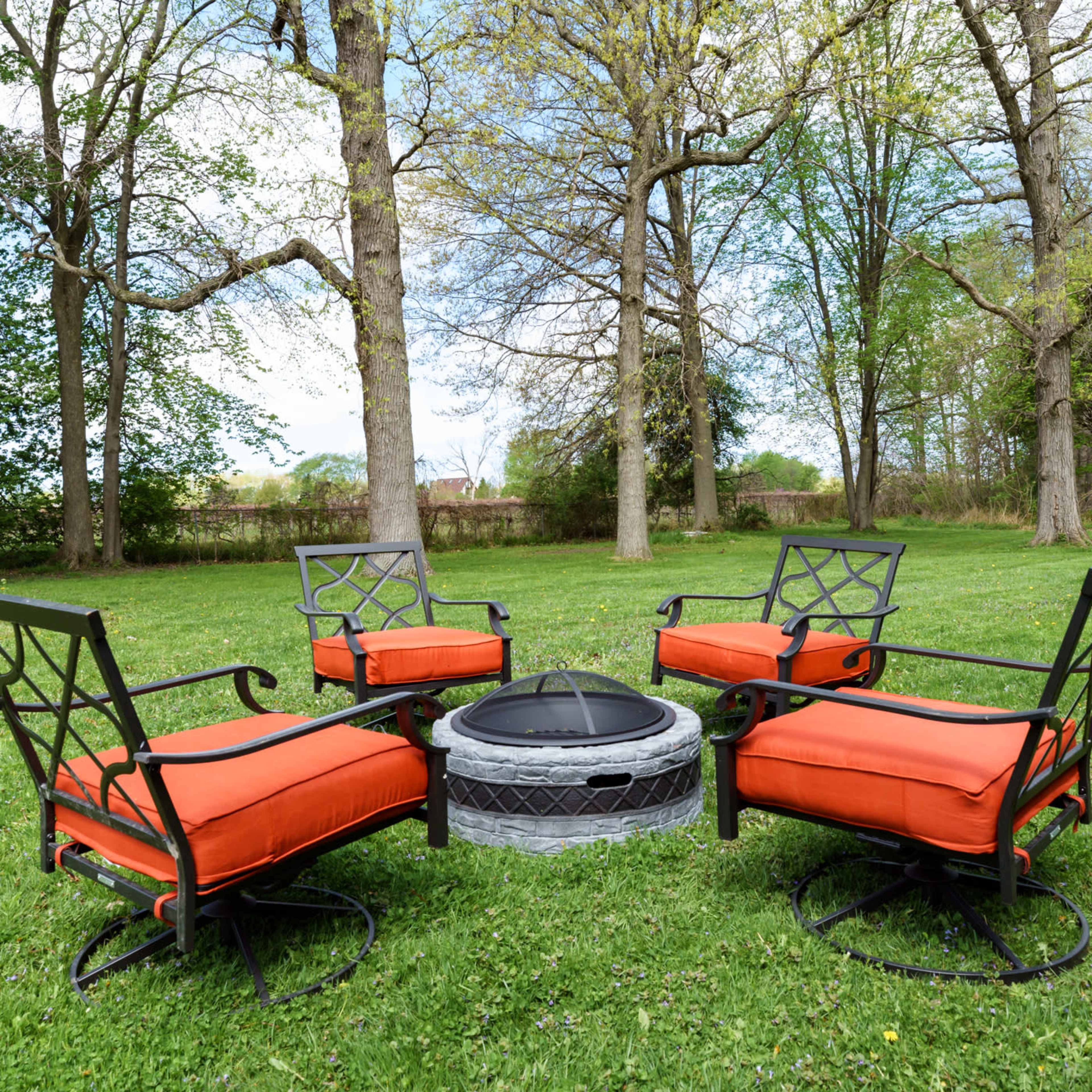 A circular fire pit is surrounded by four metal chairs with orange cushions on a grassy lawn bordered by trees.