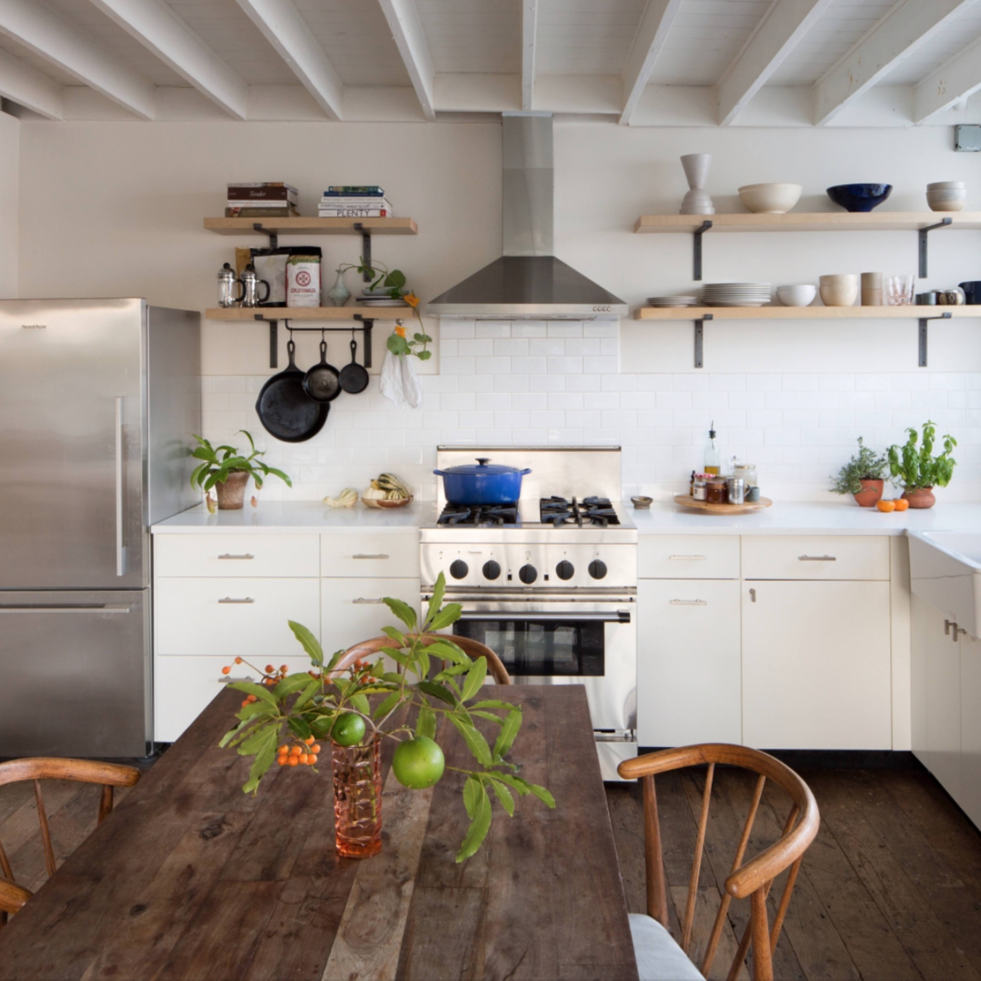 A modern kitchen features a stainless steel refrigerator, white cabinets, a gas stove with a hood, open shelving, and a wooden dining table adorned with a vase of plants.