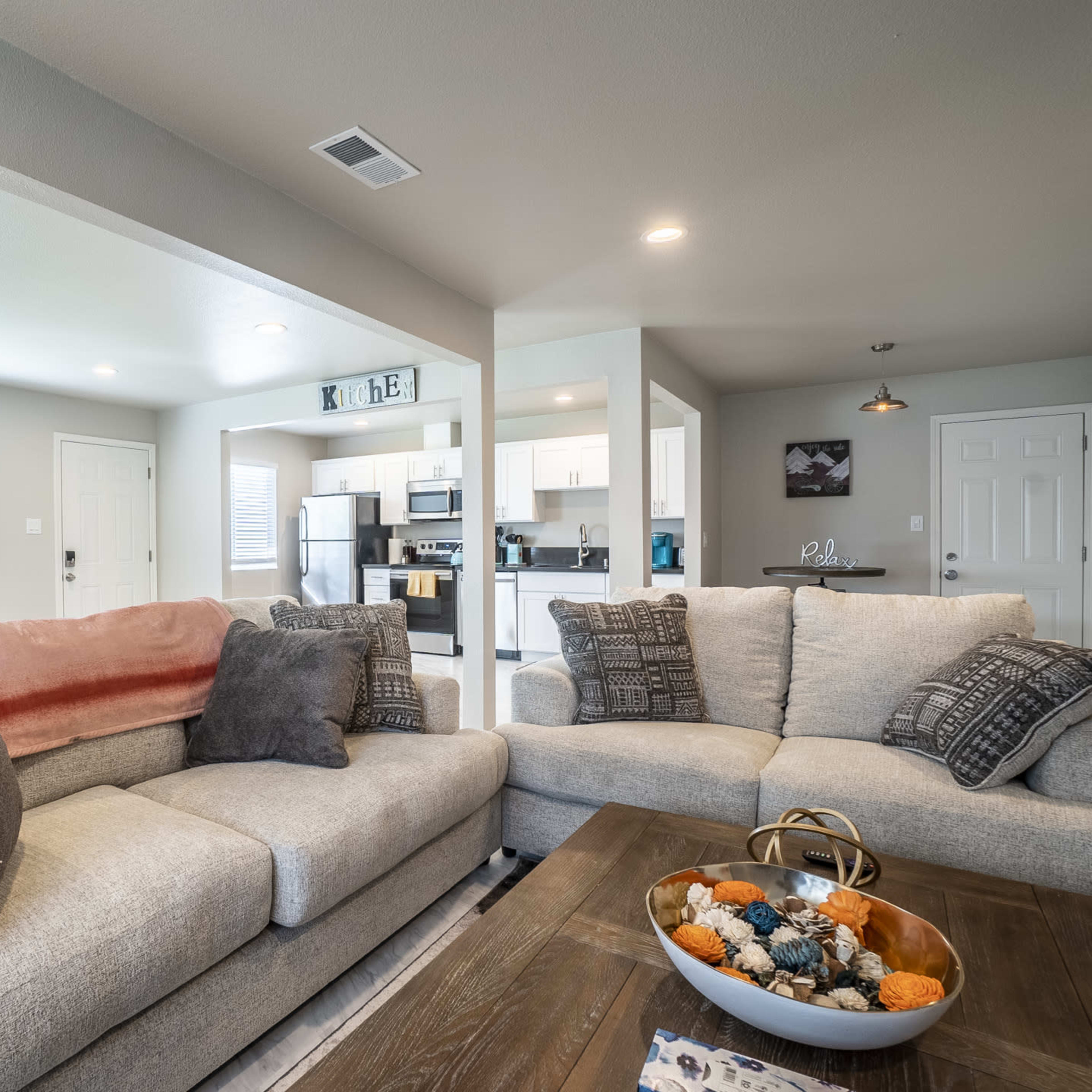 A modern living room with a light gray sofa, a wooden coffee table adorned with decorative items, and an open kitchen visible in the background.
