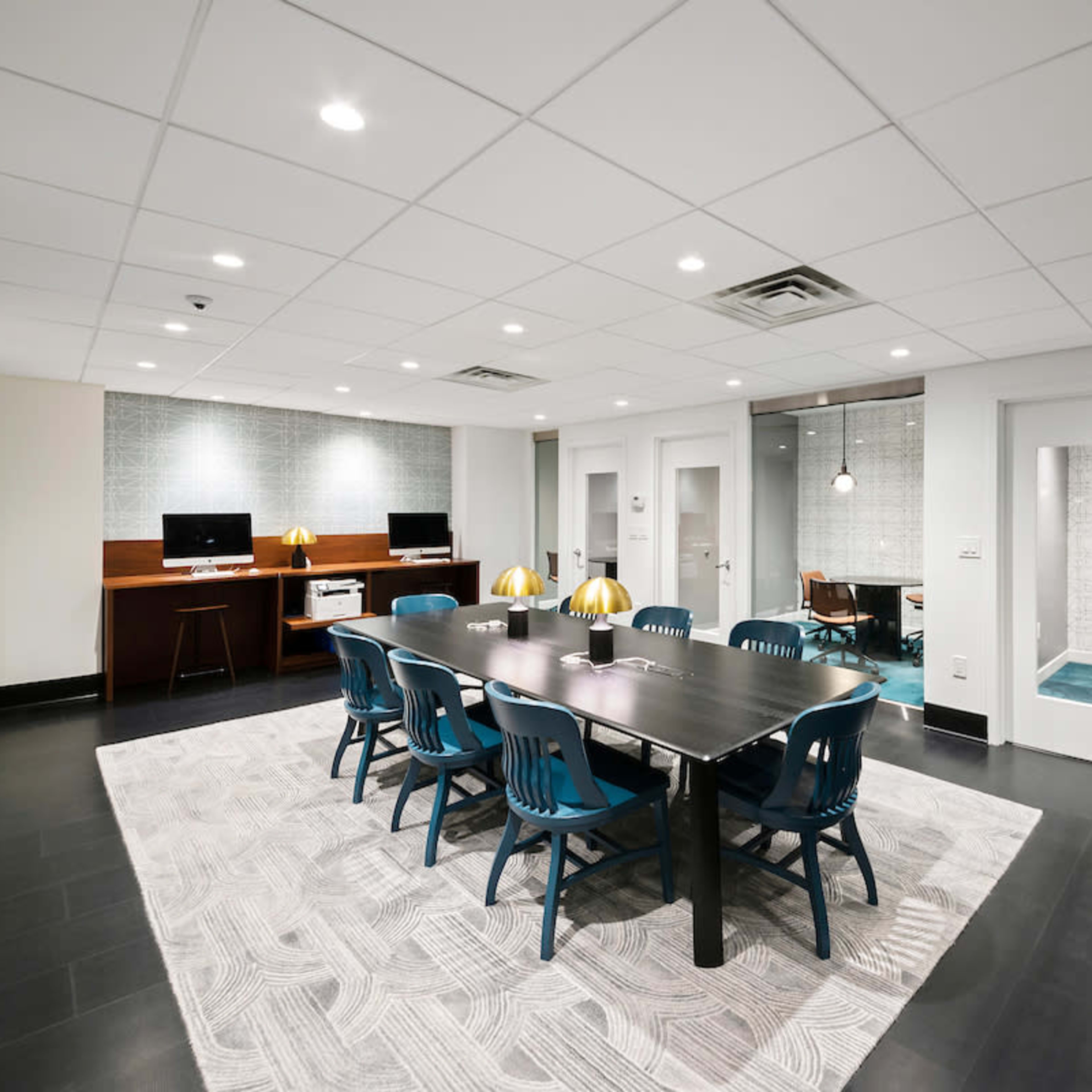 A modern conference room with a long table surrounded by blue chairs, alongside glass-walled meeting rooms and workstations.