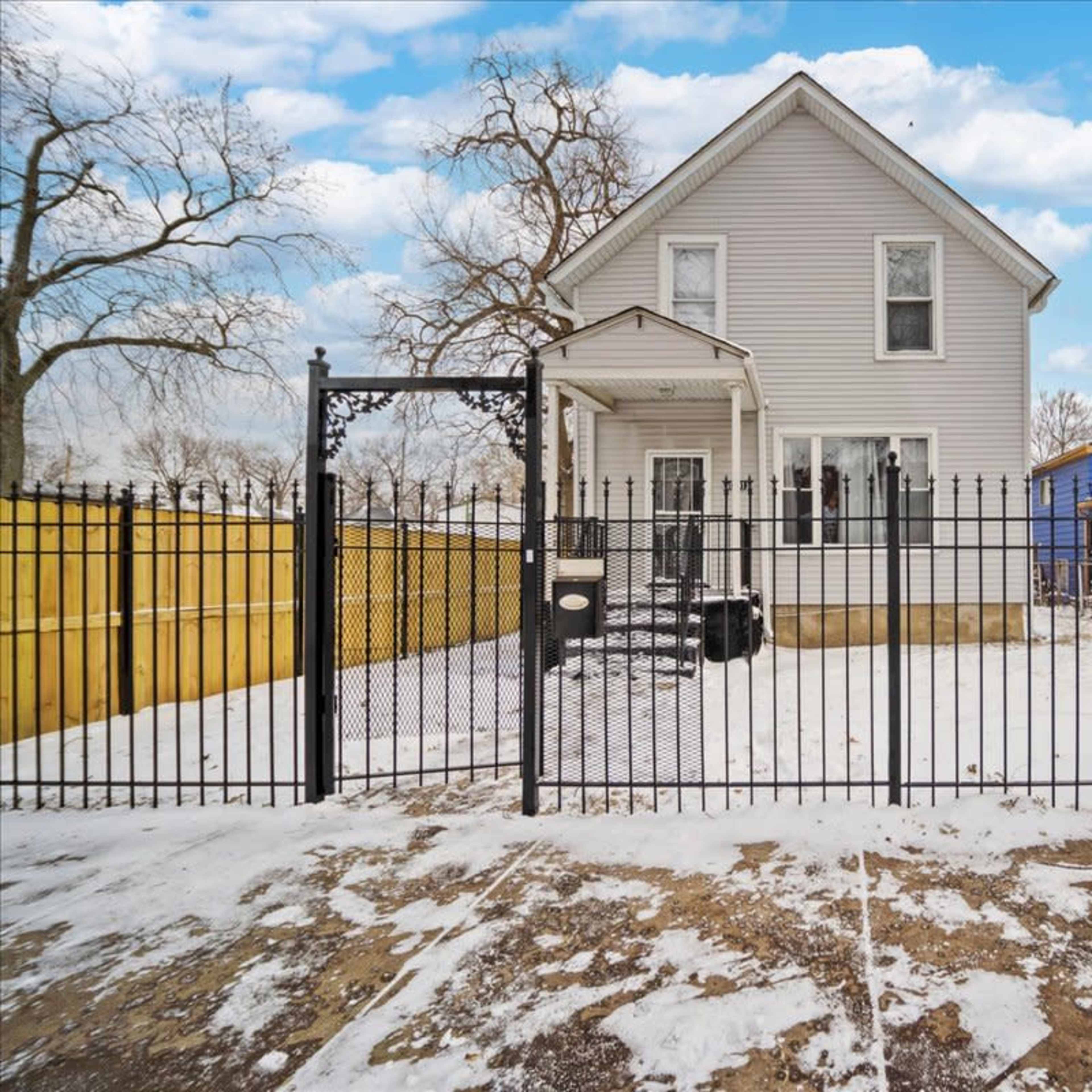A two-story house with a fenced yard, covered patio, and snow-covered ground is surrounded by trees and neighboring structures.