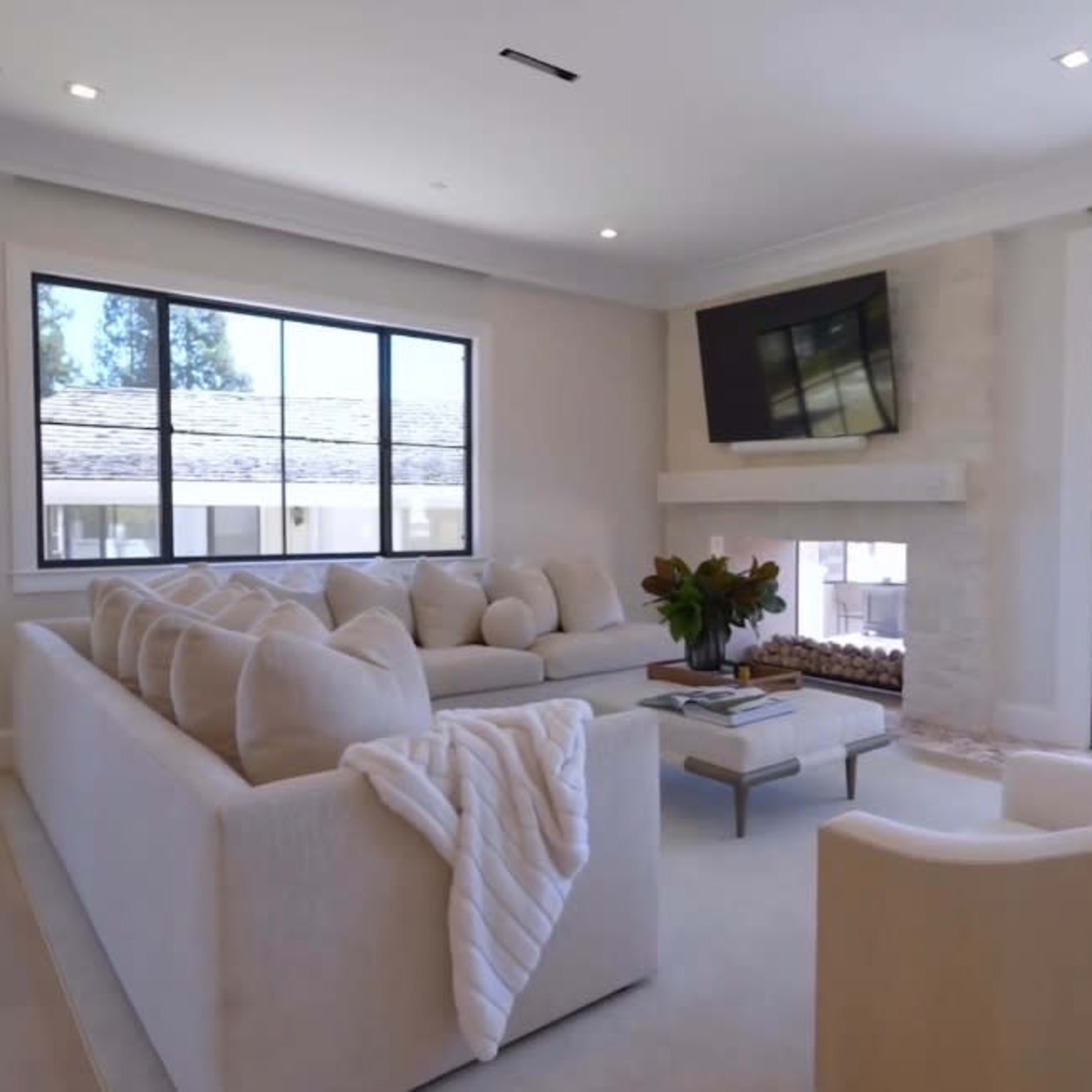 A modern living room with a large sofa, two armchairs, a television mounted on the wall, and large windows providing natural light.