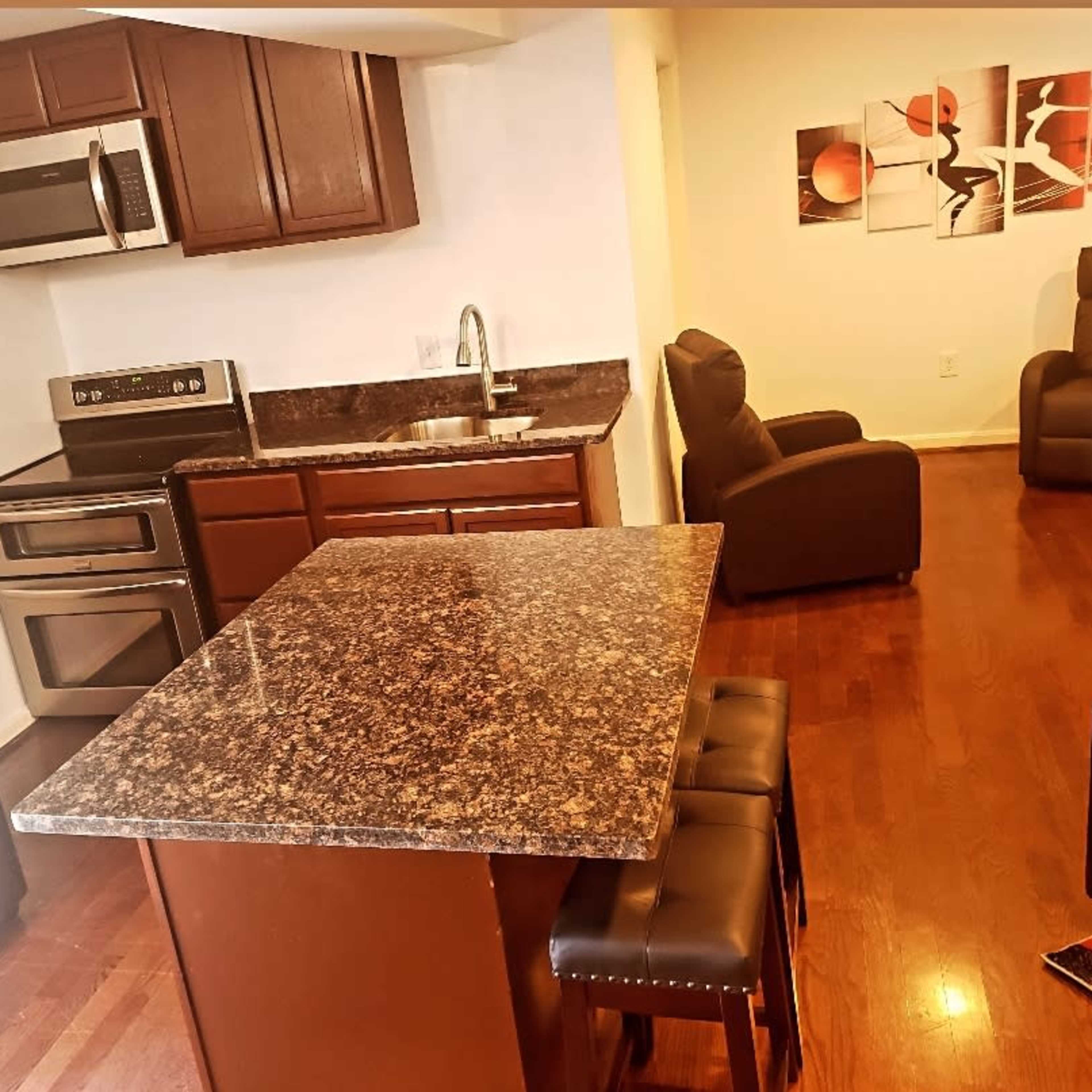 A kitchen with dark wood cabinets, stainless steel appliances, and a granite countertop island, connected to a living area with two black armchairs.