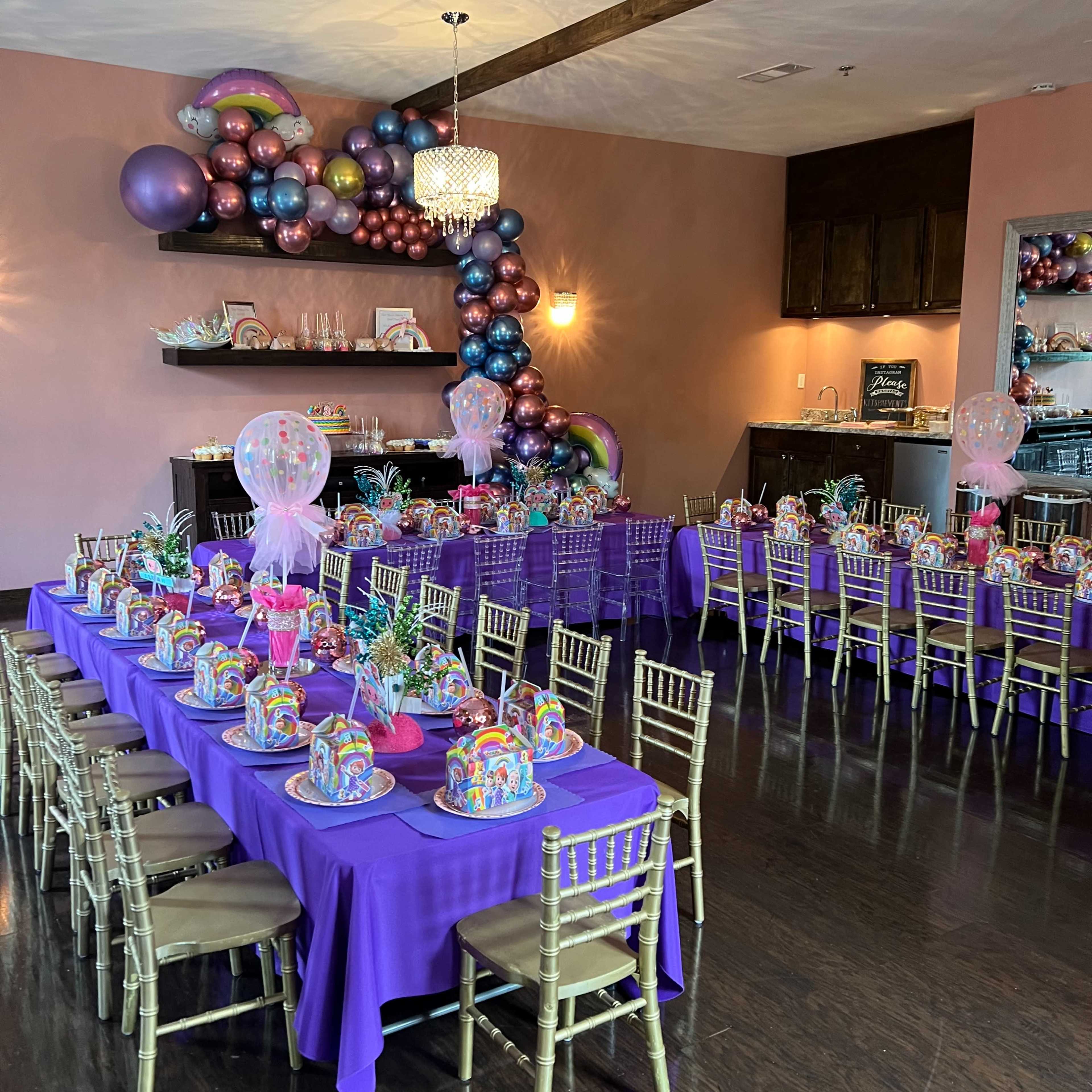 A banquet hall is set with two long tables covered in purple cloth, decorated with colorful centerpieces and balloons on one side of the room.