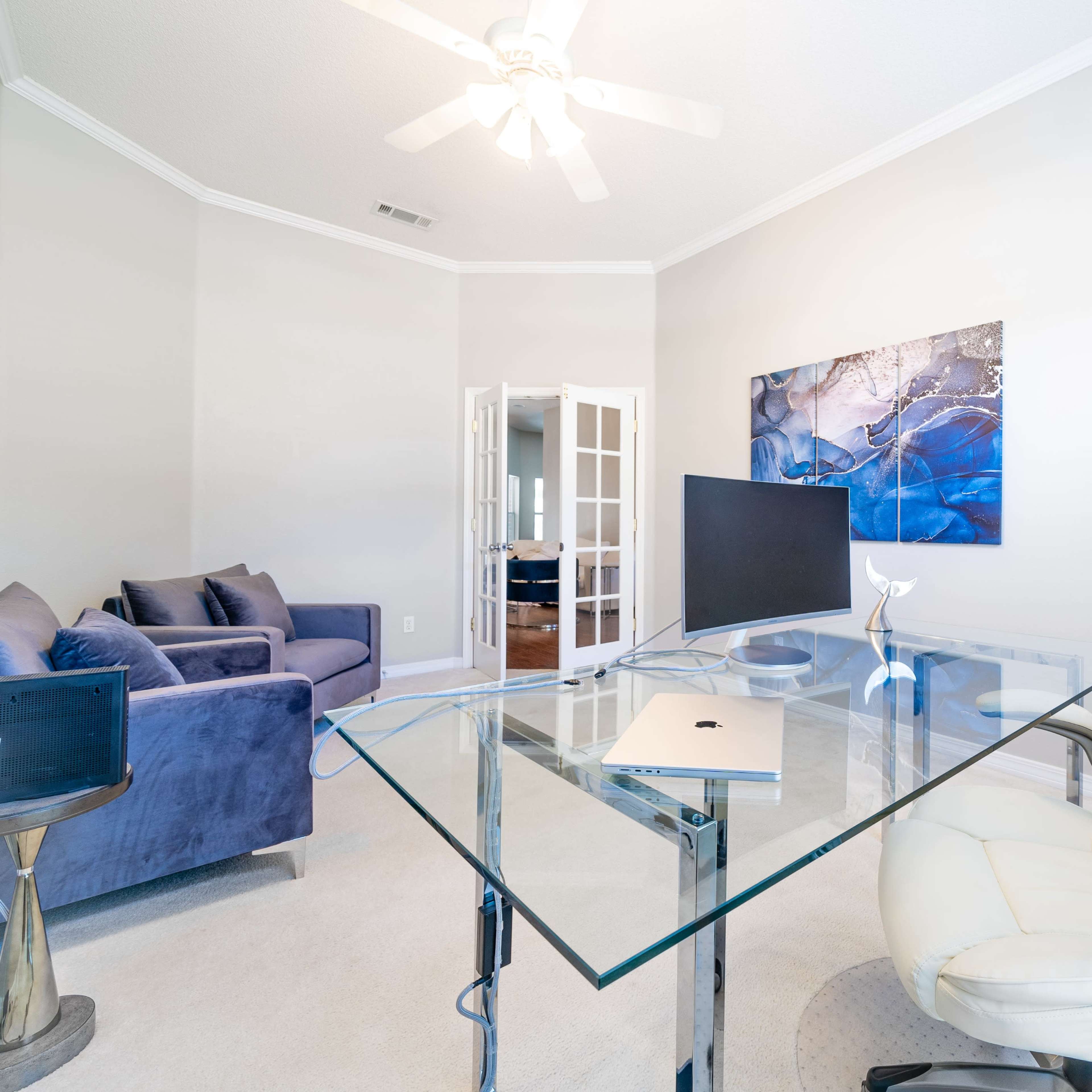 A modern office space features a clear glass desk and a white ergonomic chair, with a blue couch in the corner and a painting on the wall.