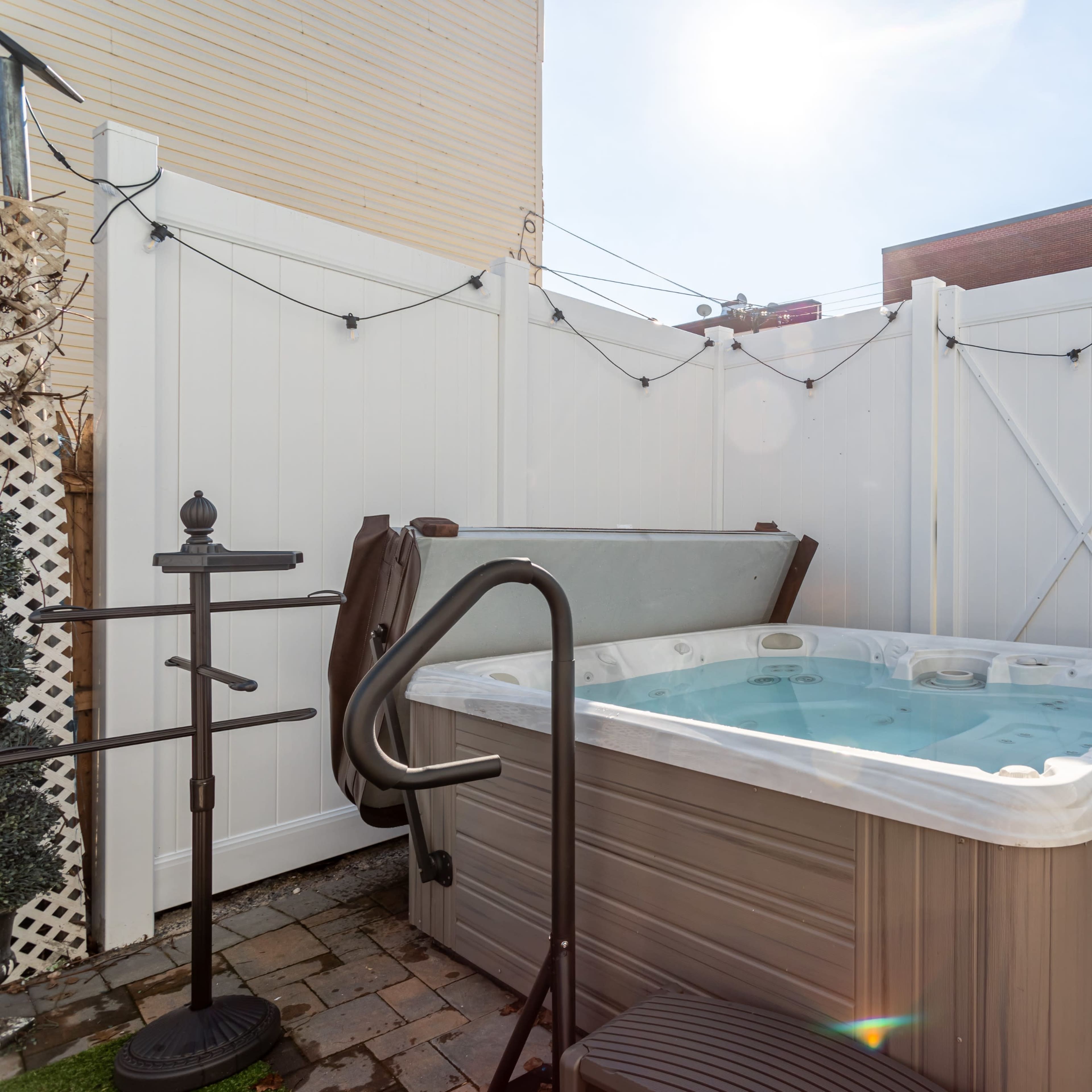 A hot tub is positioned in a small, enclosed outdoor space surrounded by white fencing and a paved floor.