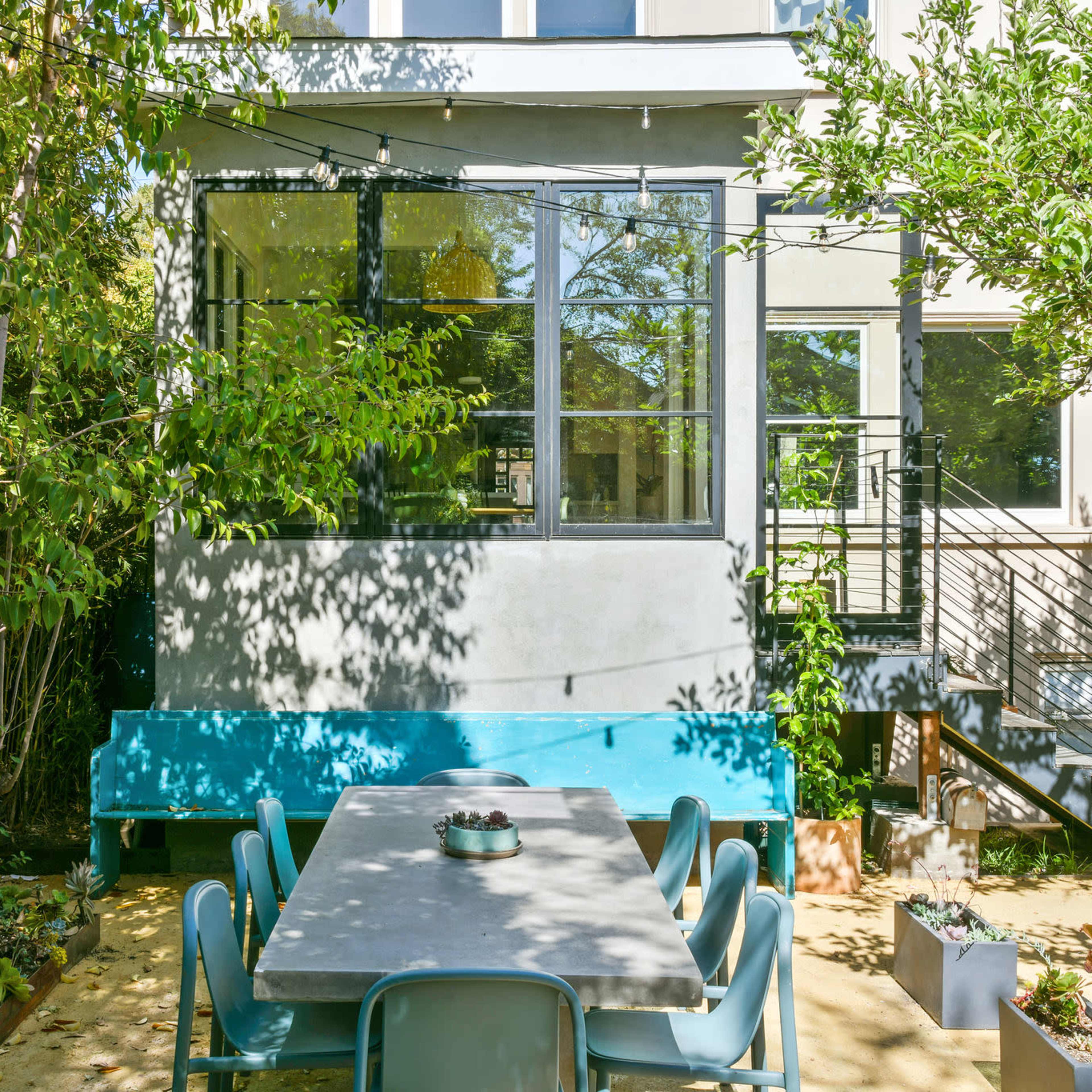 A large outdoor dining table is positioned in a garden area next to a residential building with large windows and a staircase.