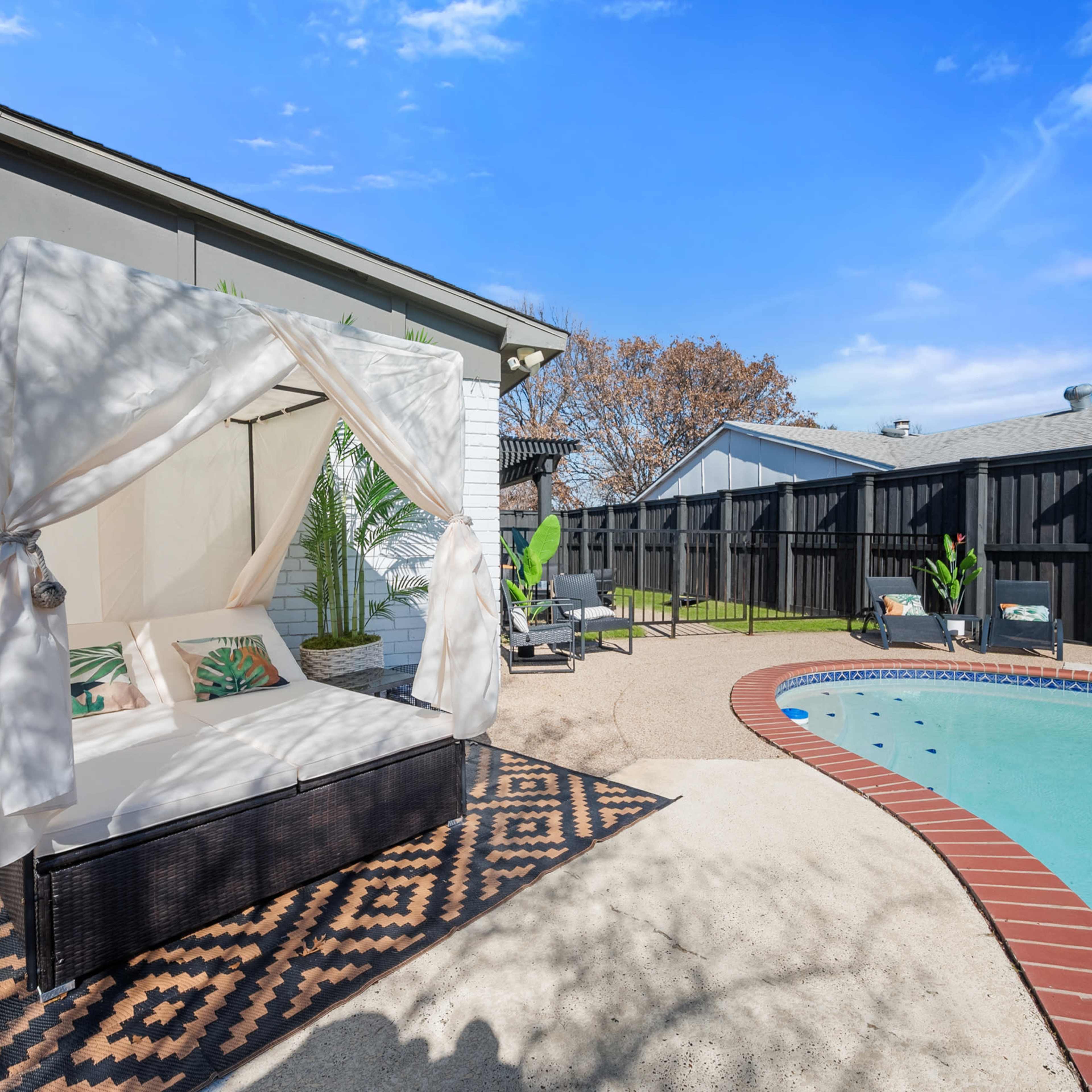 A backyard pool area featuring a covered seating space and a well-maintained swimming pool surrounded by greenery.