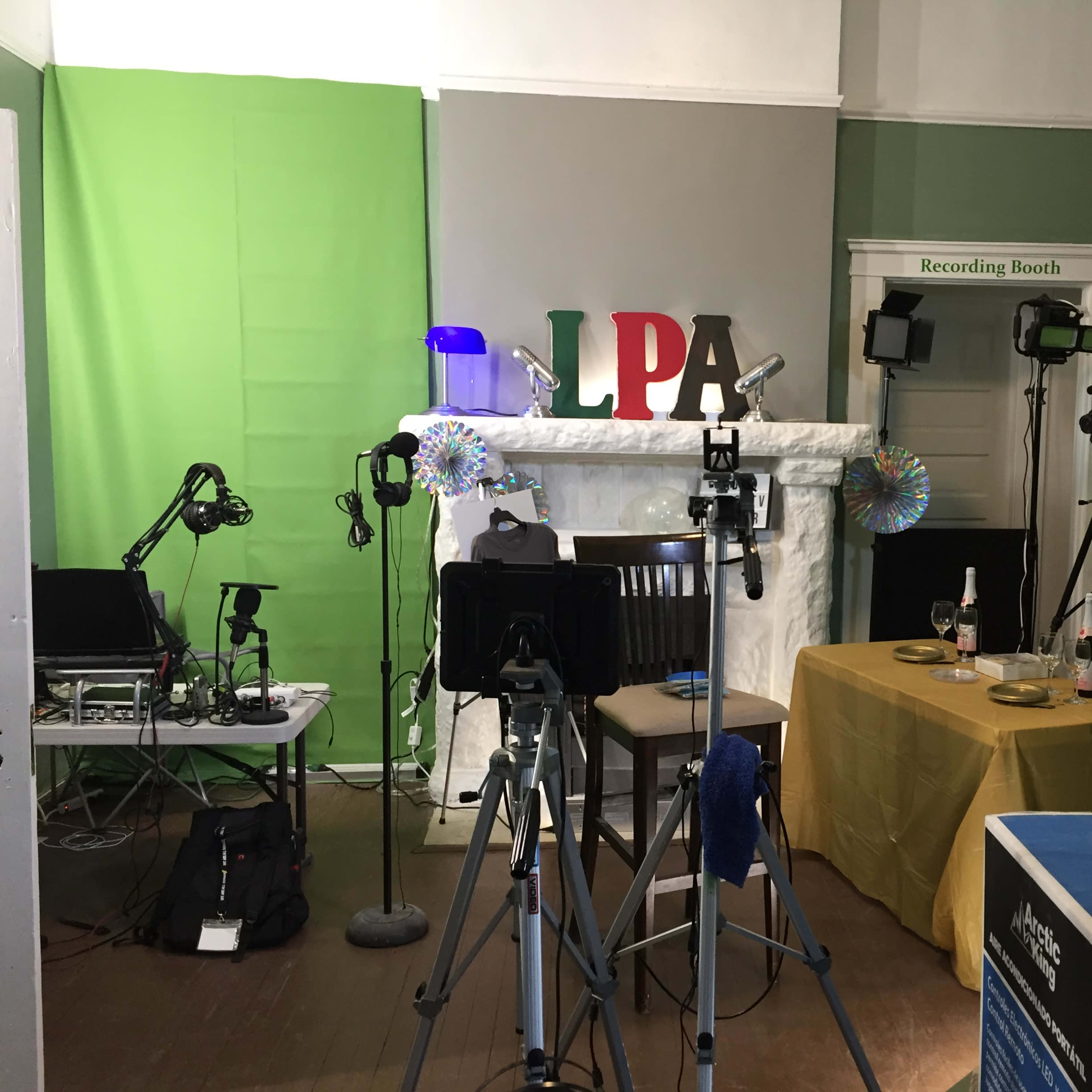 A recording studio setup with green screens, various cameras, microphones, and a table with a tablecloth, located in a room with a decorative fireplace.