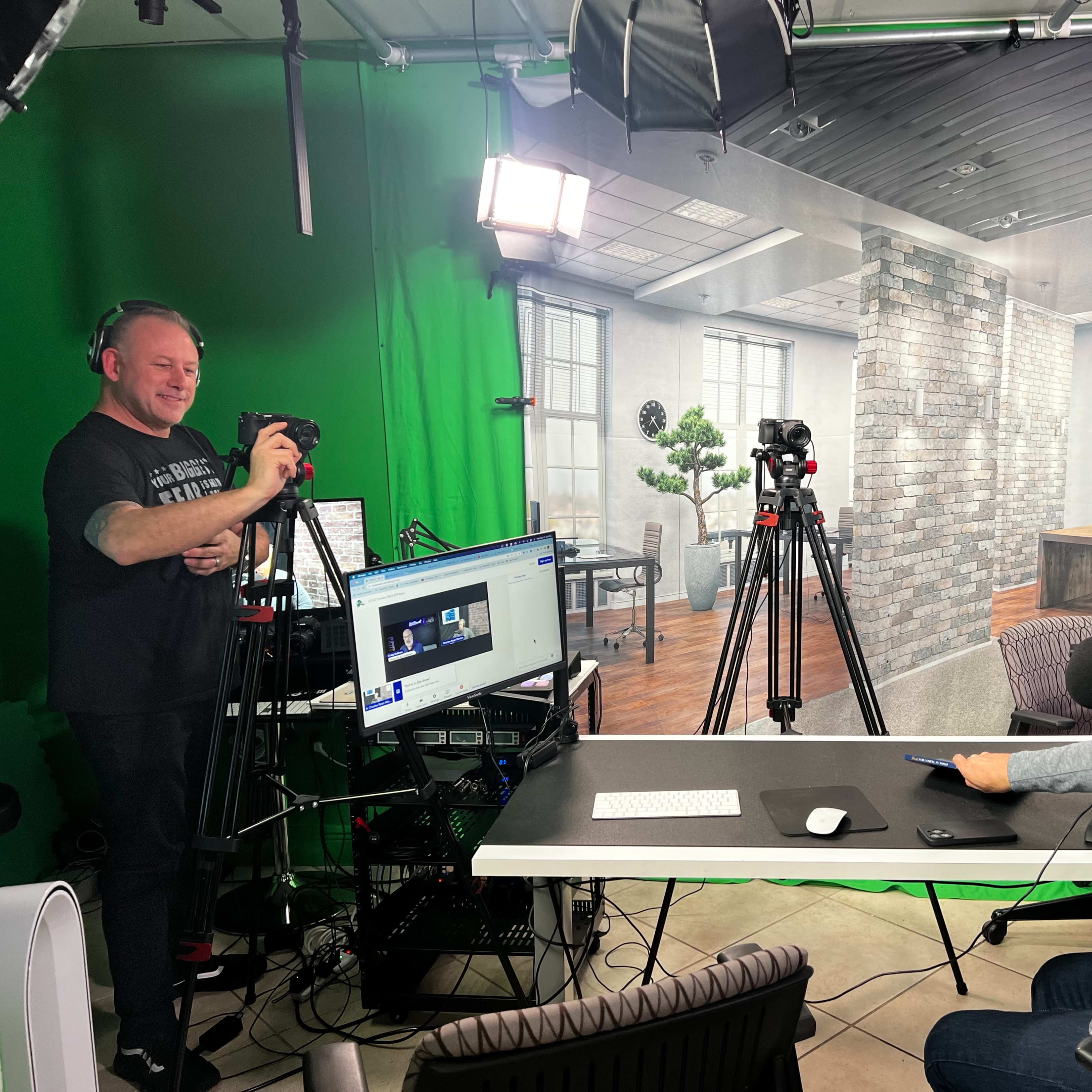 A man sits at a desk with headphones, while another man stands behind him filming with a camera in a studio setting.