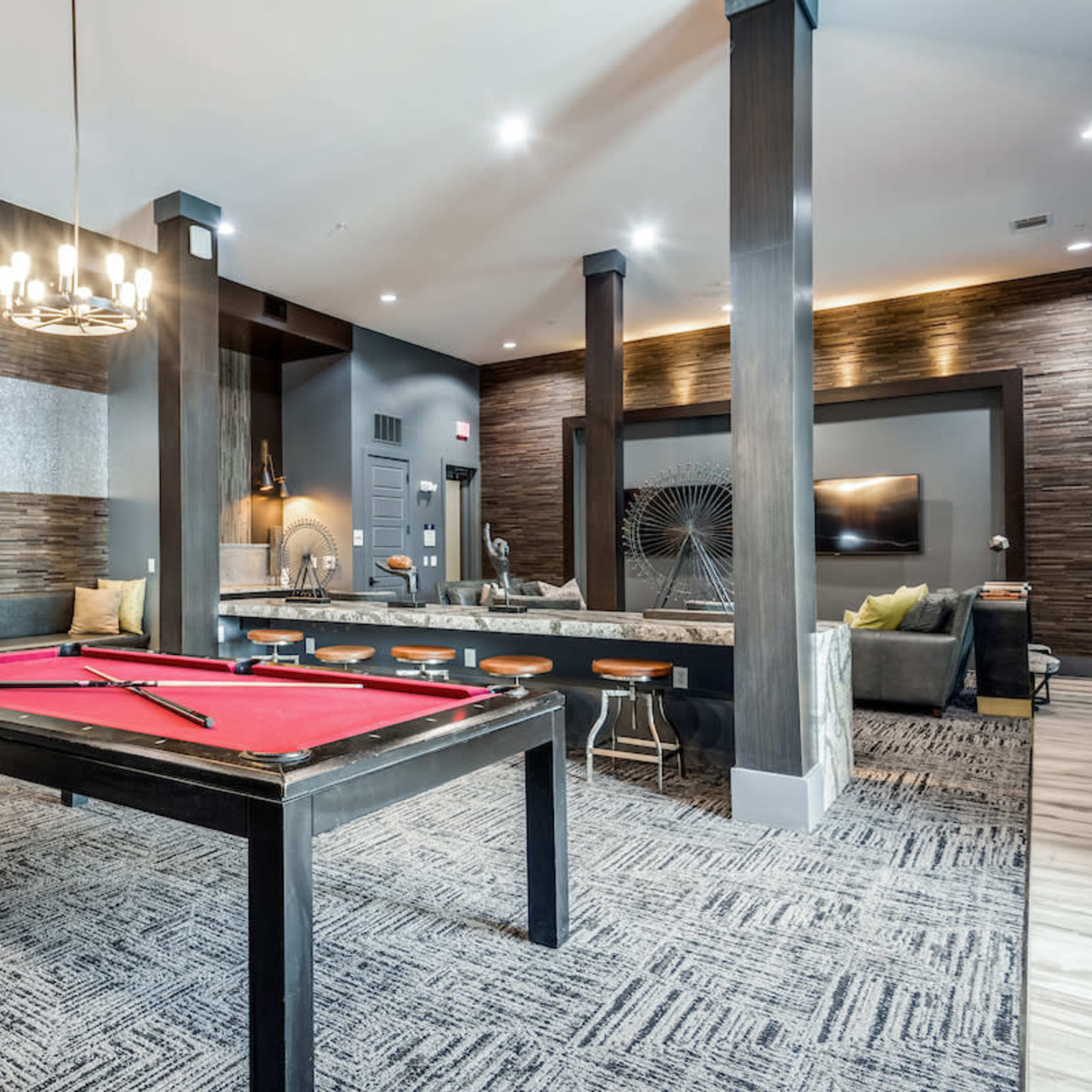 A modern lounge area with a red pool table, bar stools, and contemporary furniture set against a stylish backdrop of textured wall panels and pendant lighting.