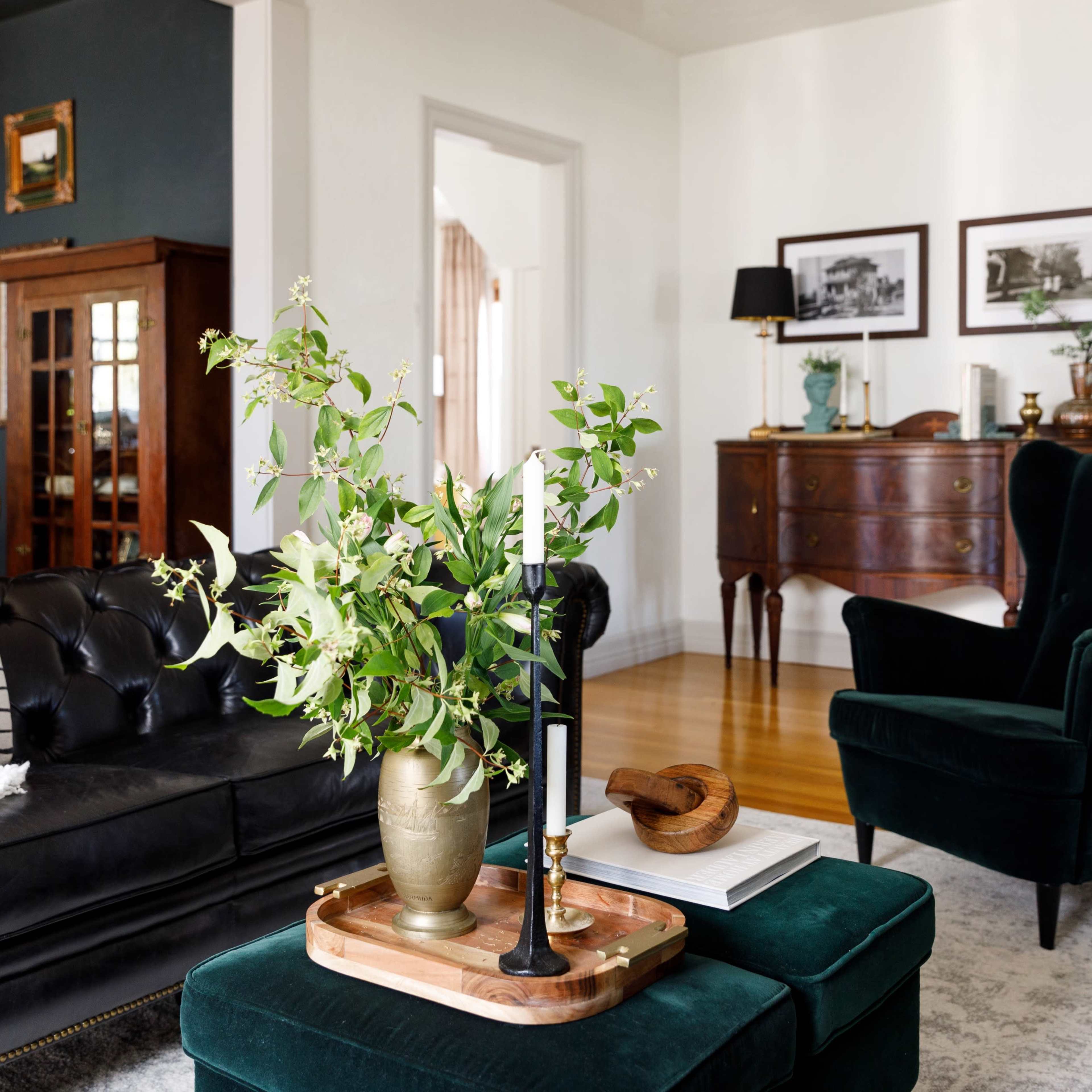 The living room features dark walls and a mix of vintage furniture, including a tufted black sofa, green armchairs, and a wooden sideboard, complemented by decorative plants and framed photographs.