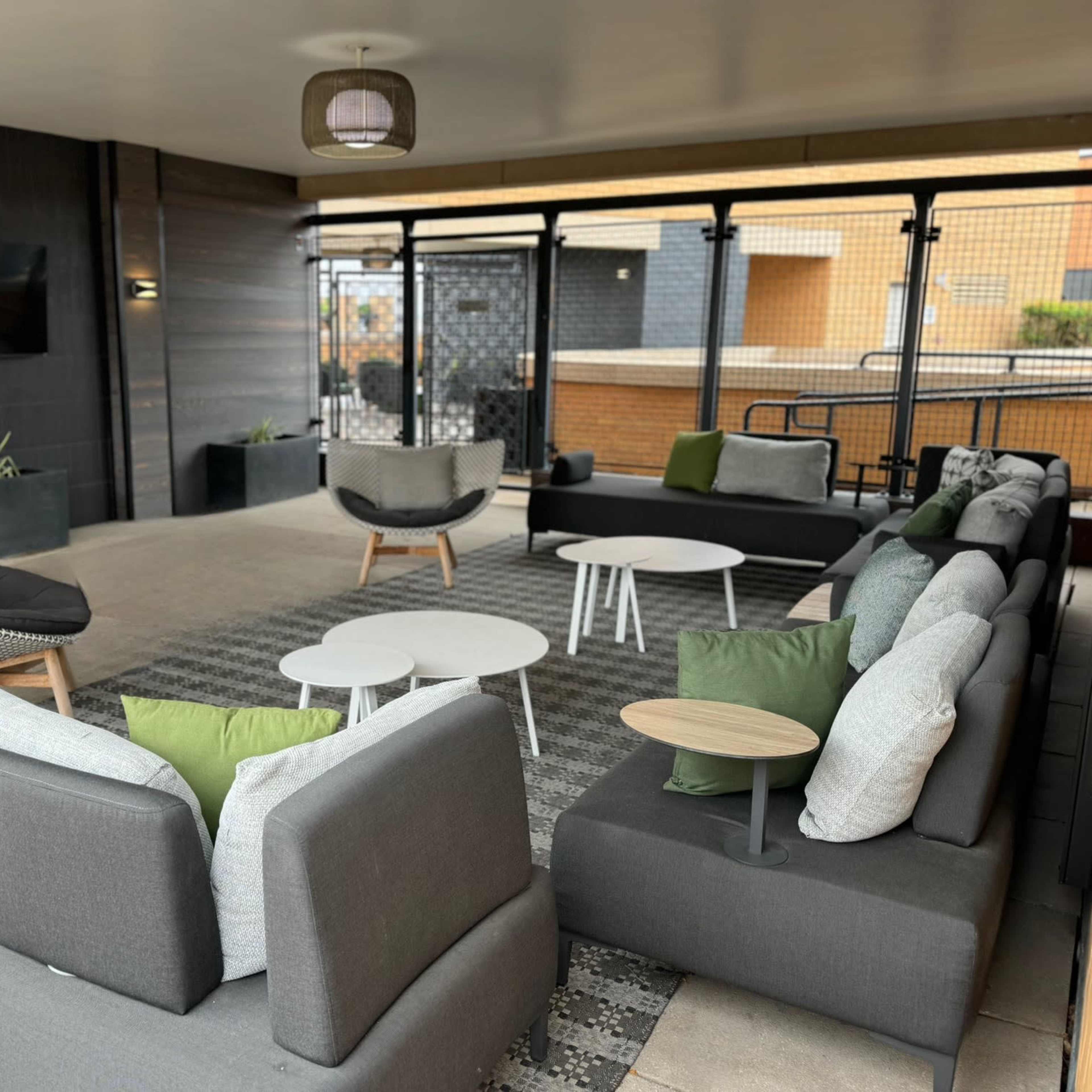 A modern lounge area with multiple gray sofas, round tables, and green accent pillows, enclosed by a glass wall.