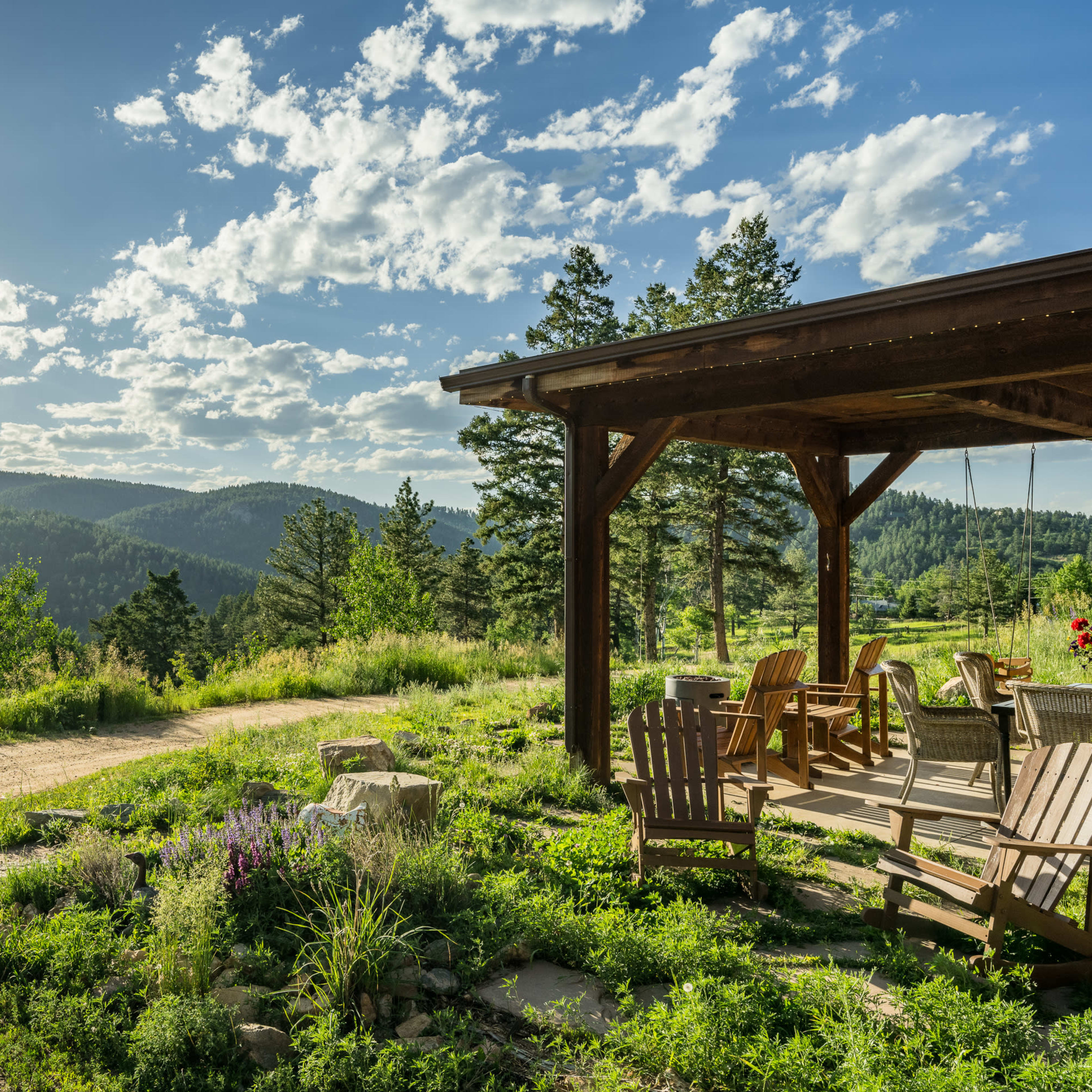 A wooden porch with chairs overlooks a green landscape of rolling hills and scattered trees under a blue sky with white clouds.