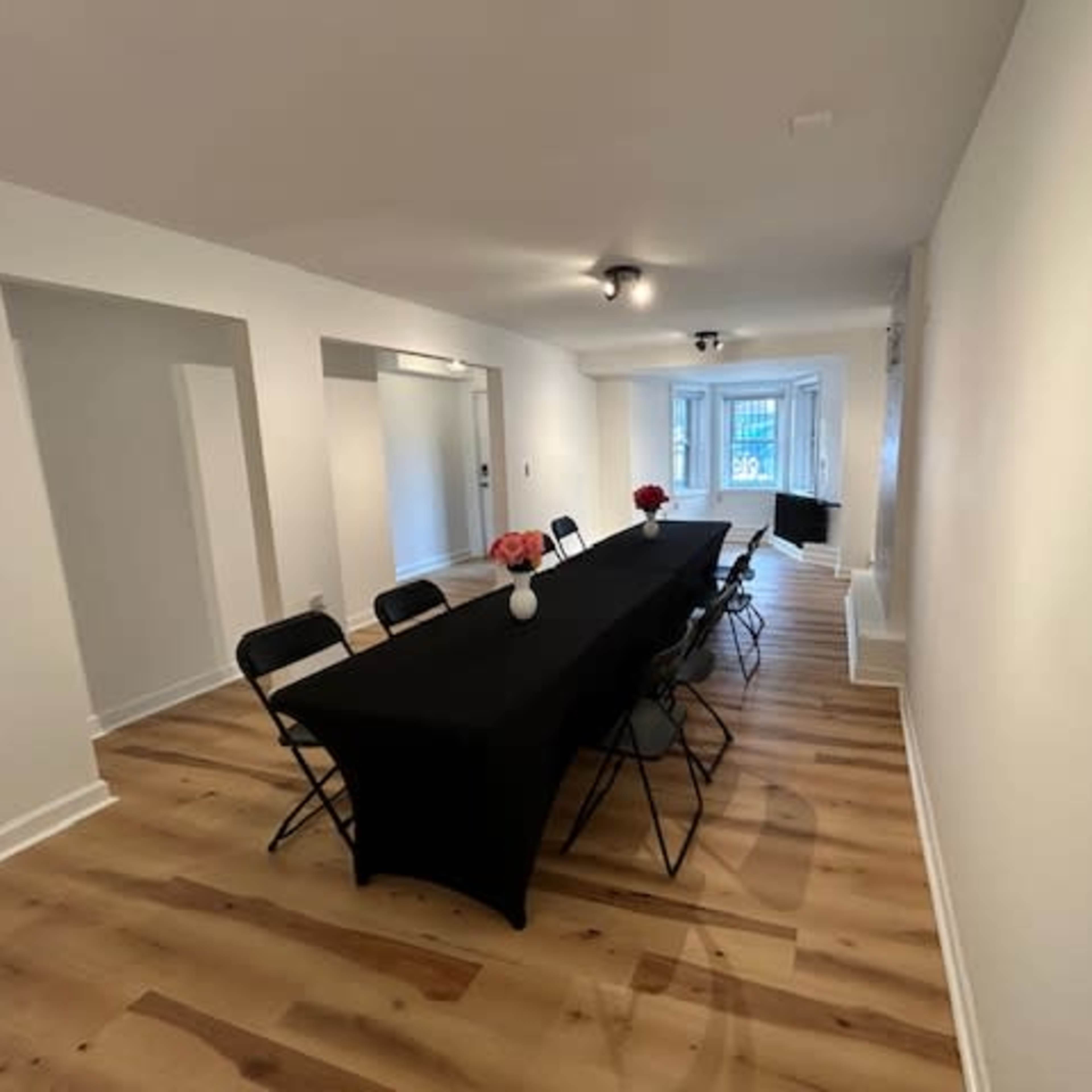 A long black table with chairs set in a bright, minimalist room featuring wooden flooring and a window at the far end.