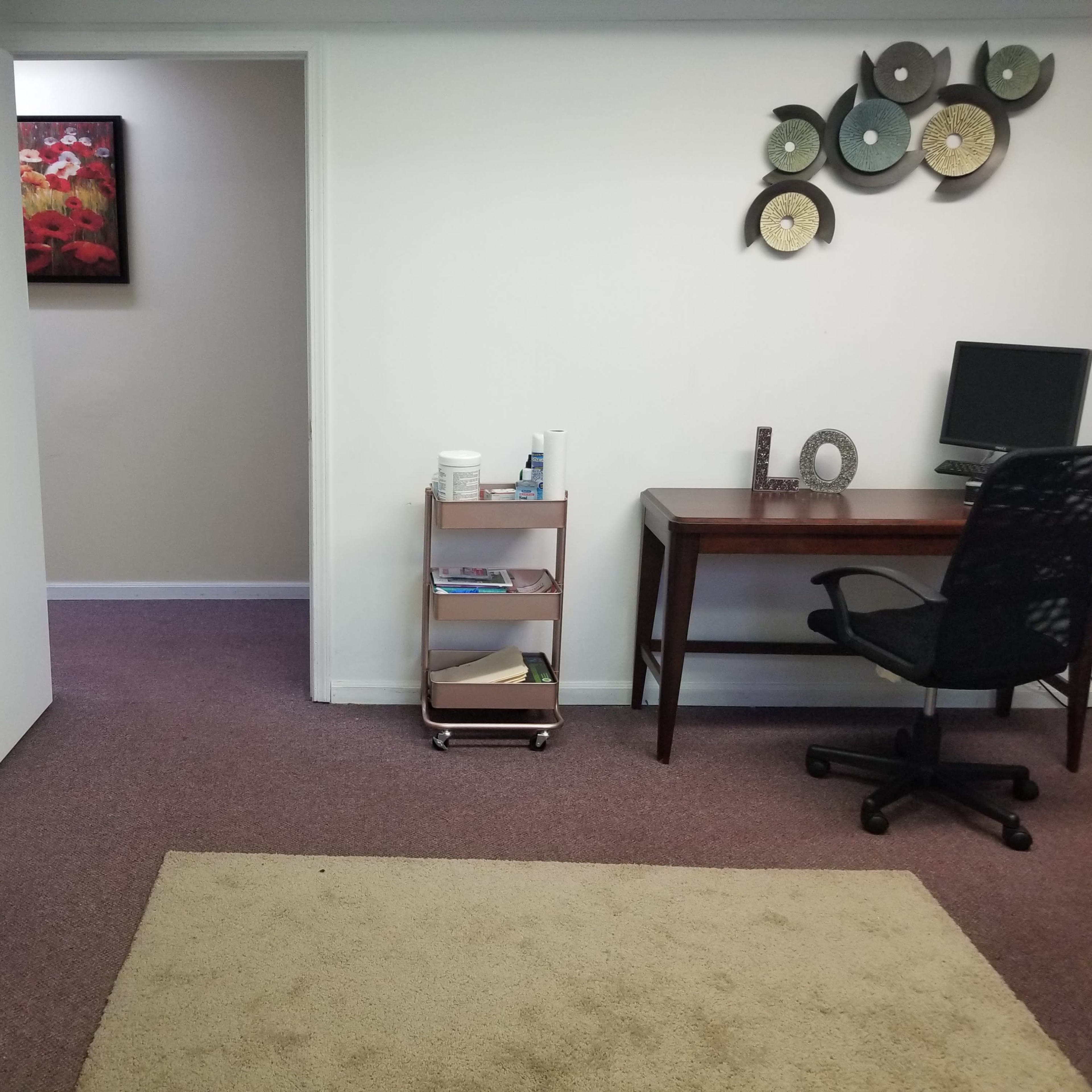 A small office space with a desk and chair, a bookshelf, and a doorway leading to another room, decorated with a wall hanging and a plant.