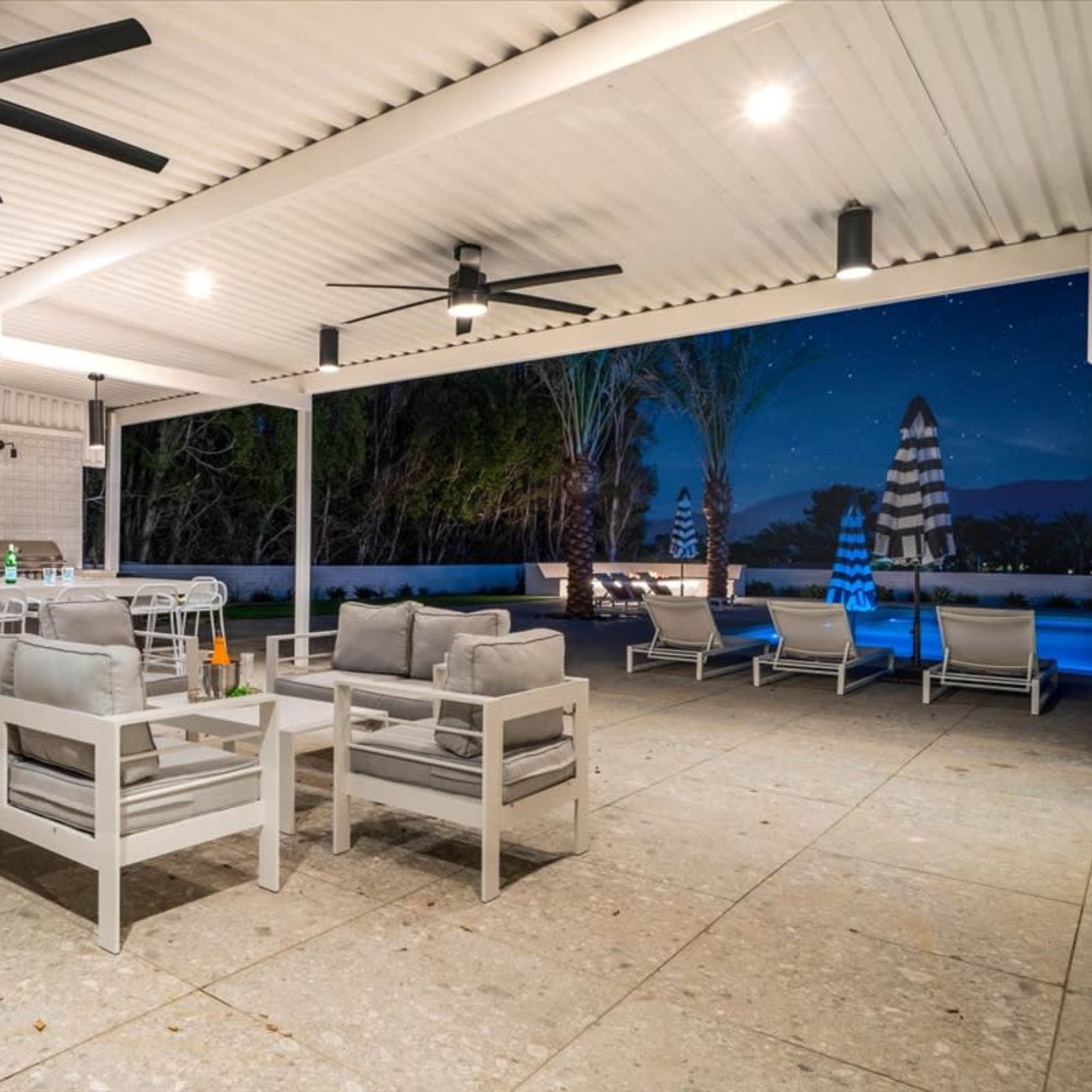 An outdoor patio area with seating, an outdoor kitchen, and lounge chairs beside a pool under a night sky.