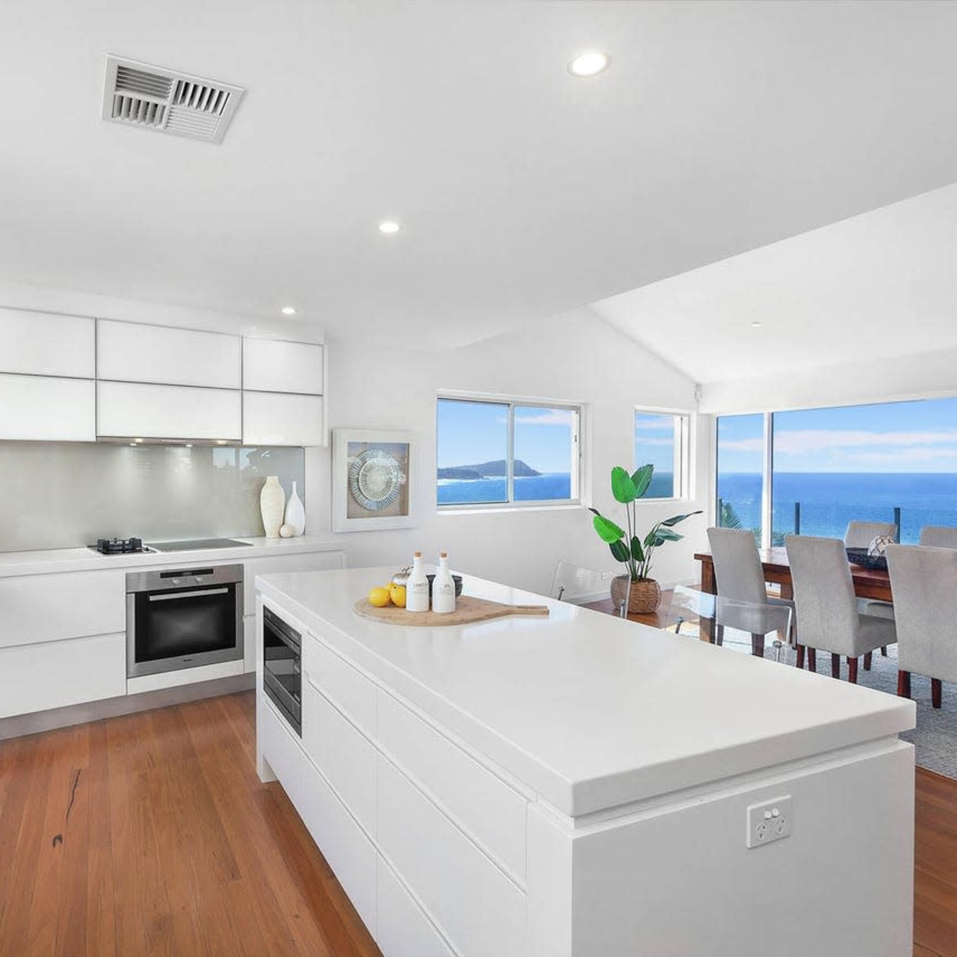 A modern kitchen with white cabinetry, a central island, and large windows overlooking a dining area with ocean views.