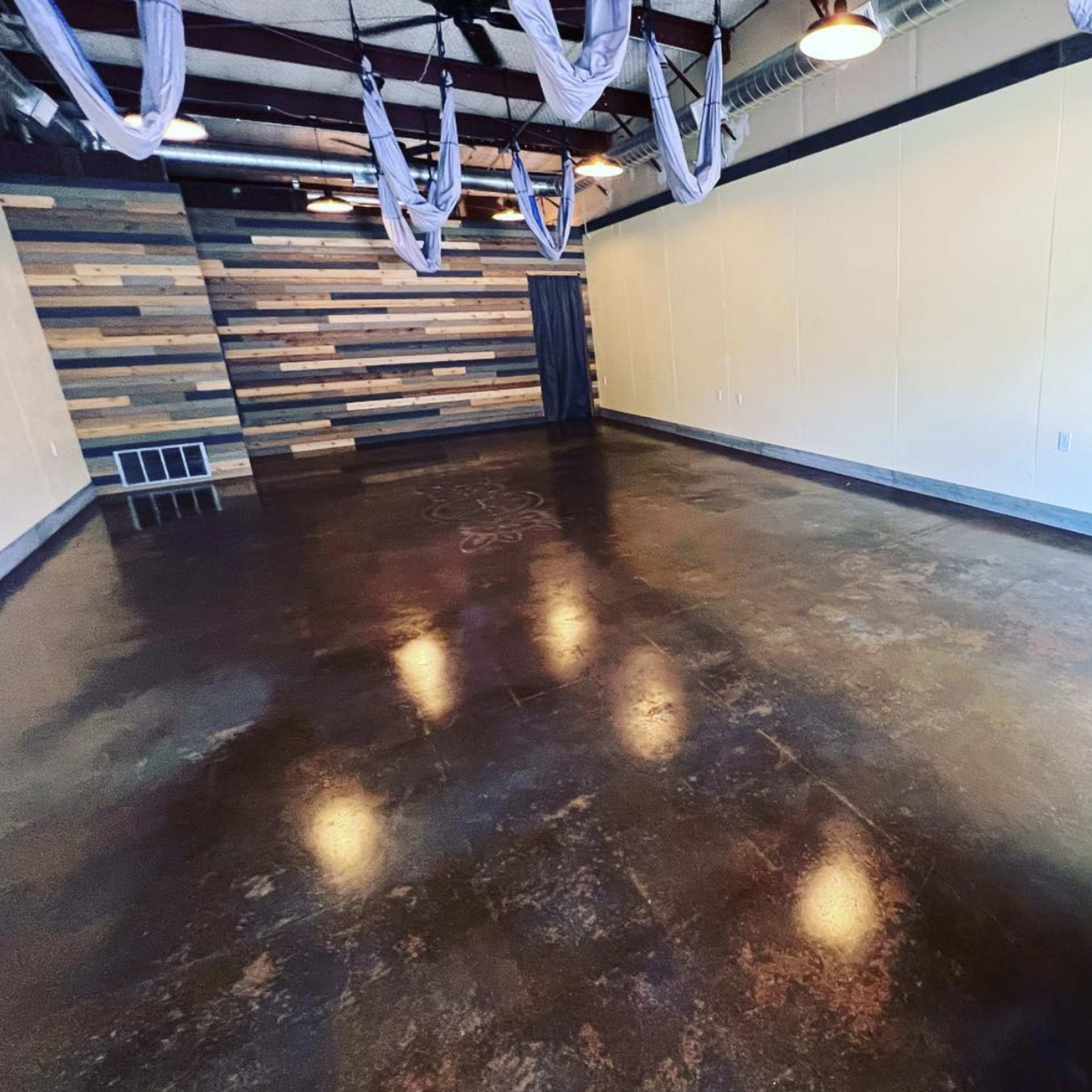An empty room with a polished concrete floor, wooden paneling on one wall, and aerial silks hanging from the ceiling.