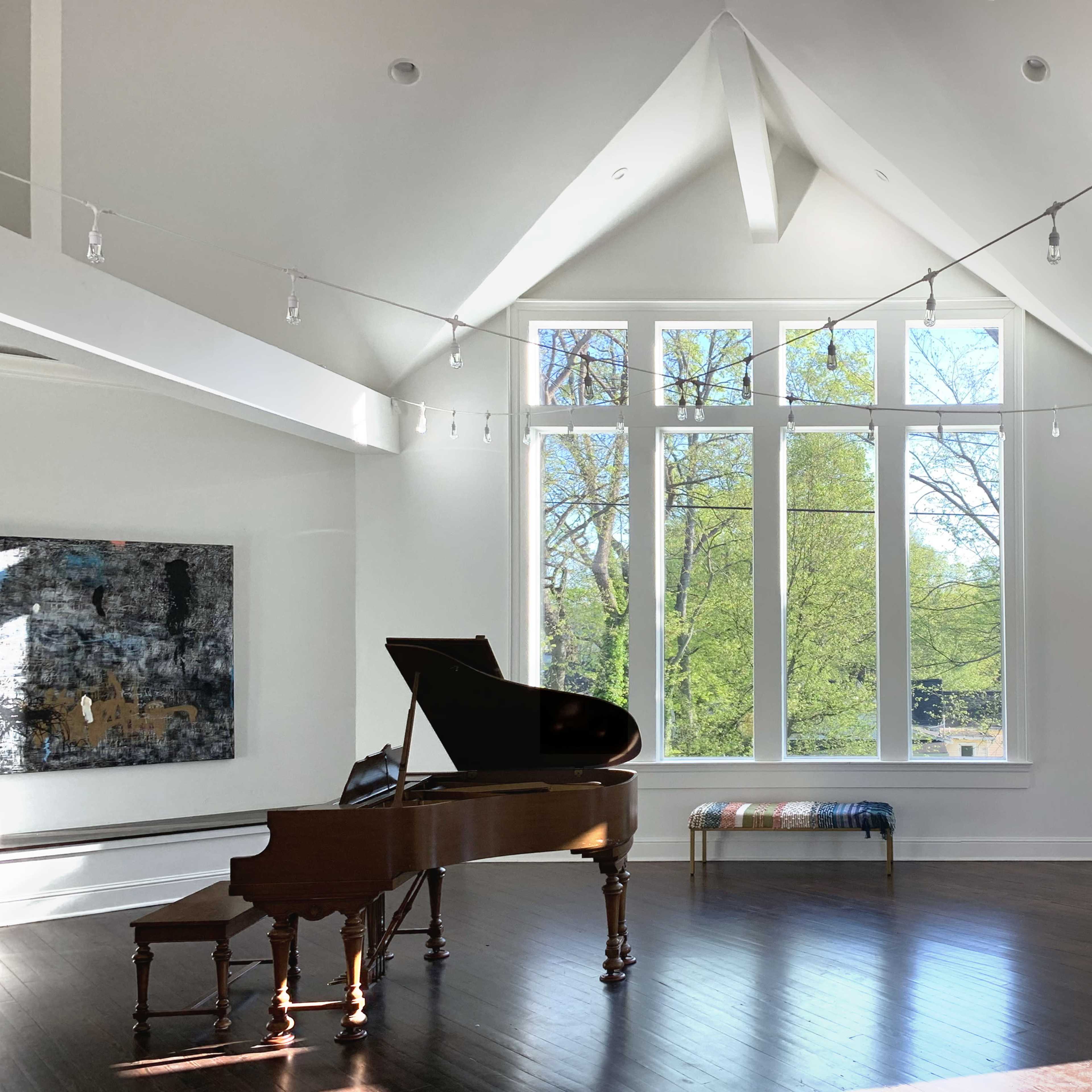 A grand piano is positioned in a brightly lit room featuring large windows that overlook greenery, with contemporary artwork on the walls and a long bench nearby.